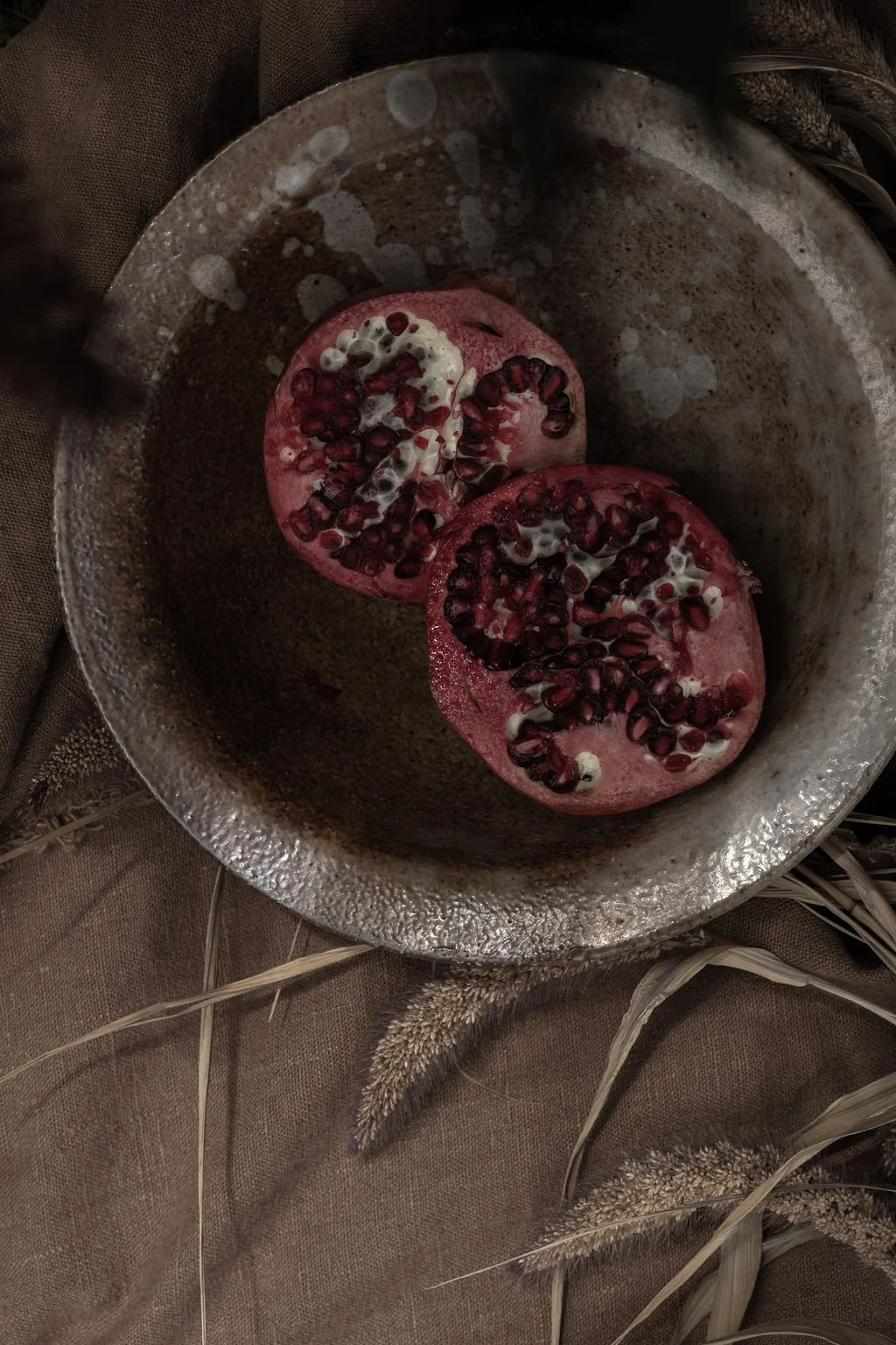 Two halves of a pomegranate in a rustic metal bowl, with seeds and white pulp visible. The bowl is placed on a brown fabric surface with some dried grass around.