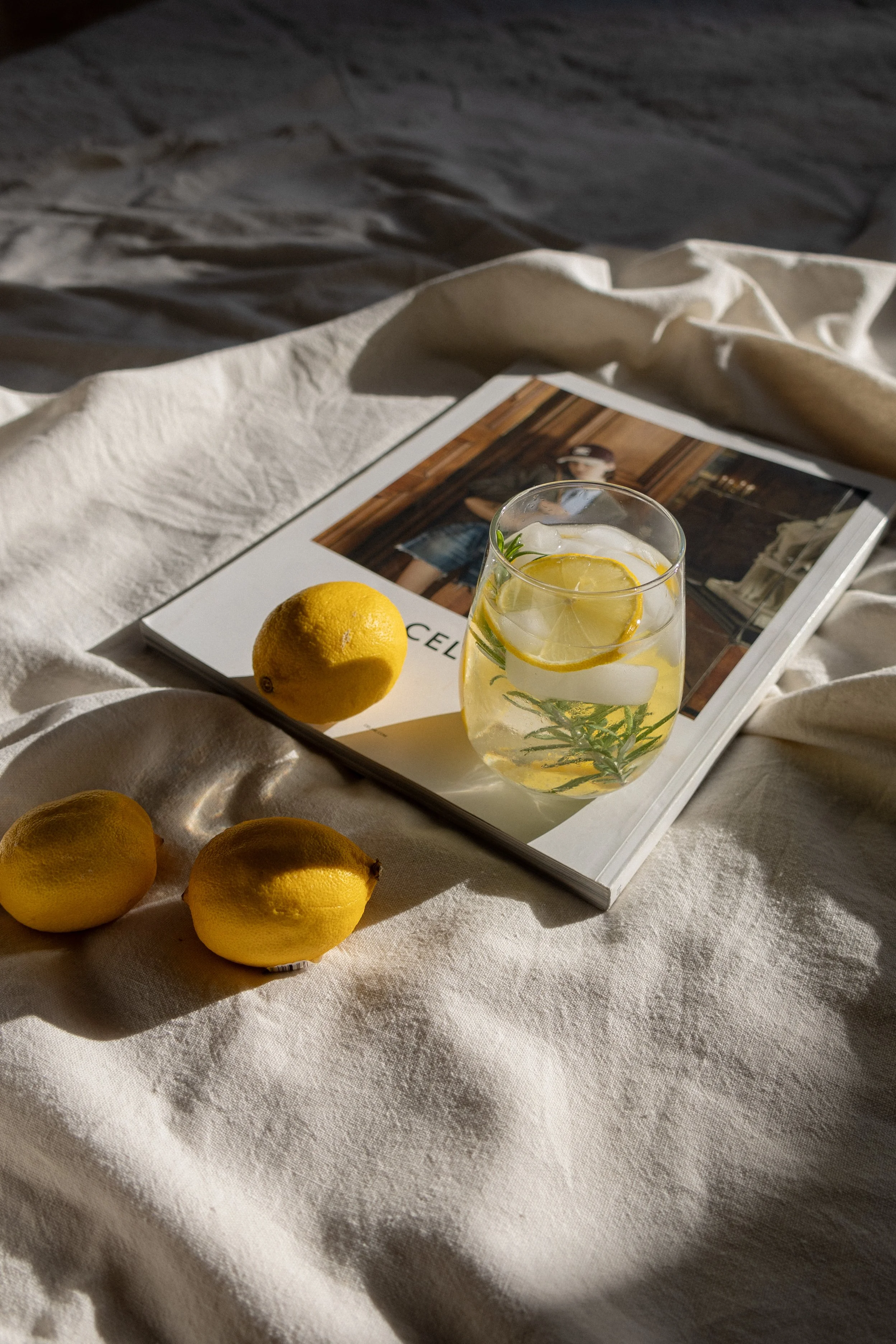 A glass of lemon and herb infused beverage on a magazine, with three lemons on a light fabric surface, illuminated by sunlight.