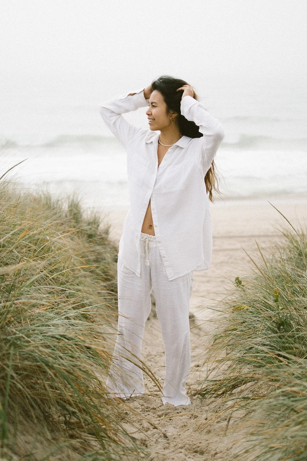 A woman standing on a sandy pathway between grass on a beach, wearing a white outfit and smiling.