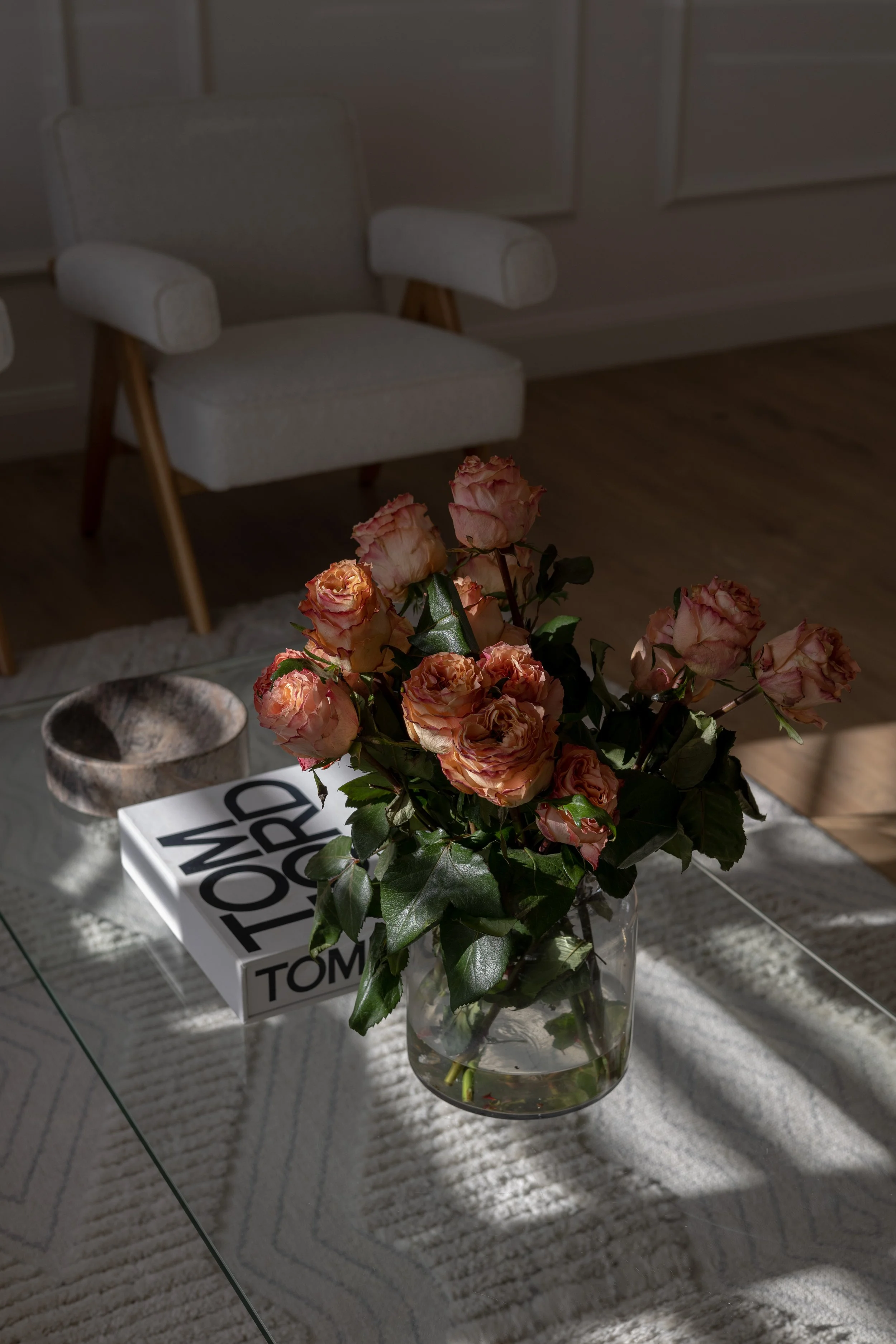 A glass vase with pink roses on a table next to a stack of books, with sunlight casting shadows on a white carpeted rug.
