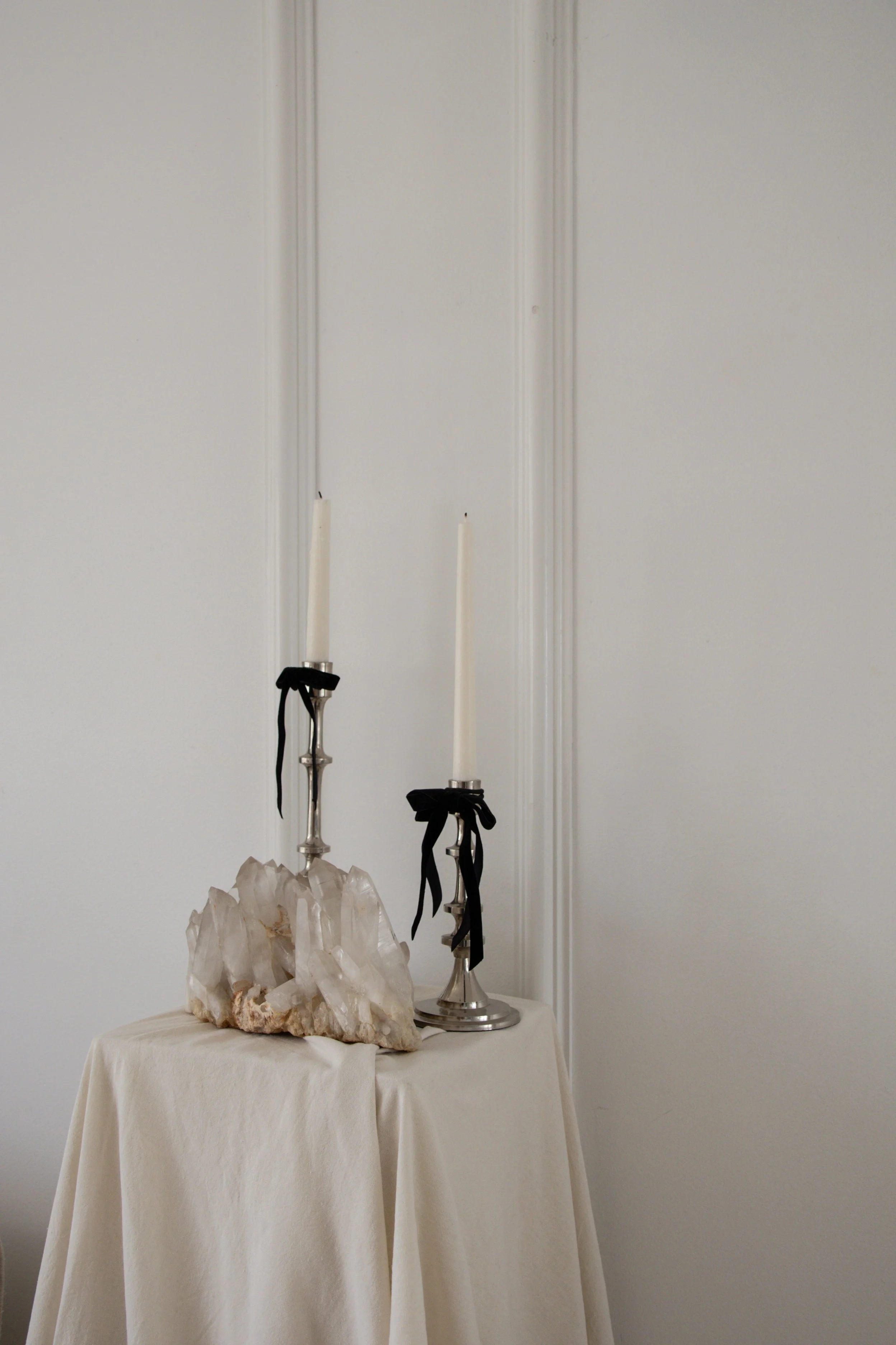 Two tall white candles with black ribbons on silver candlesticks, placed on a table covered with a cream cloth, and a large crystal formation, all against a plain white wall.