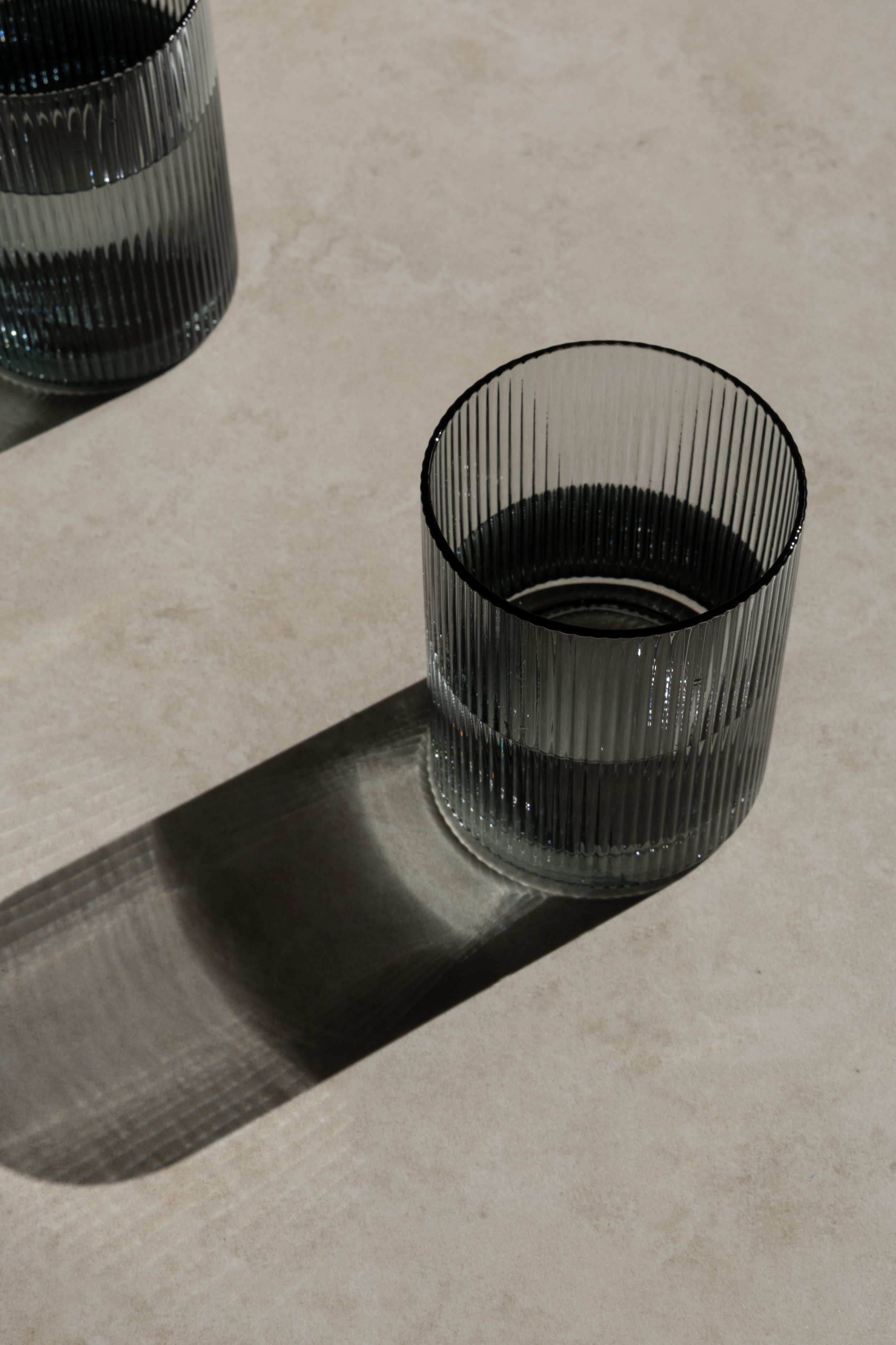 A ribbed glass tumbler with water on a beige surface, casting a shadow.
