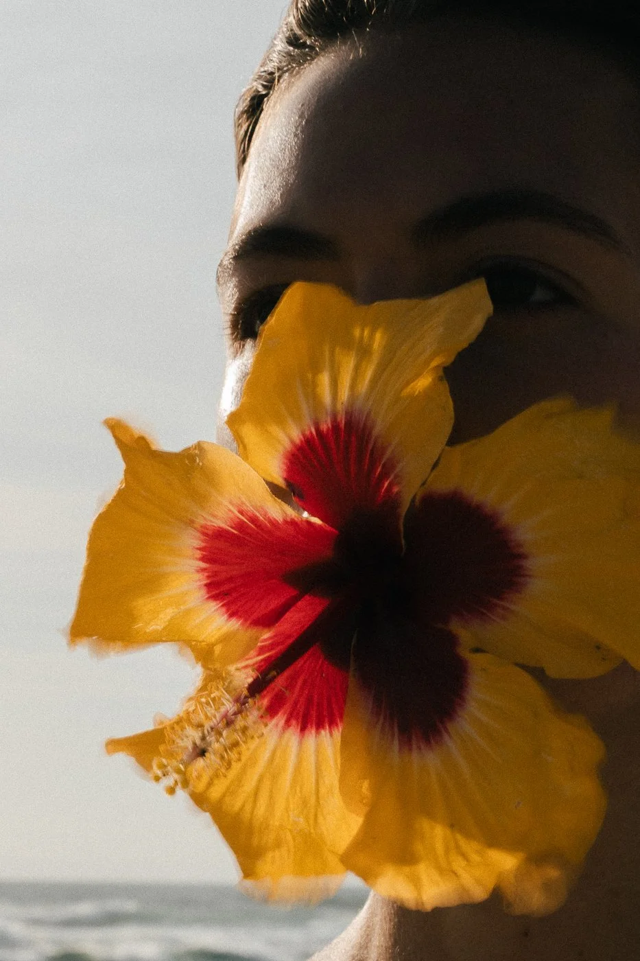 A close-up of a person's face with a large yellow Hawaiian flower with red and dark purple accents covering their nose and mouth, with a blurred outdoor background.