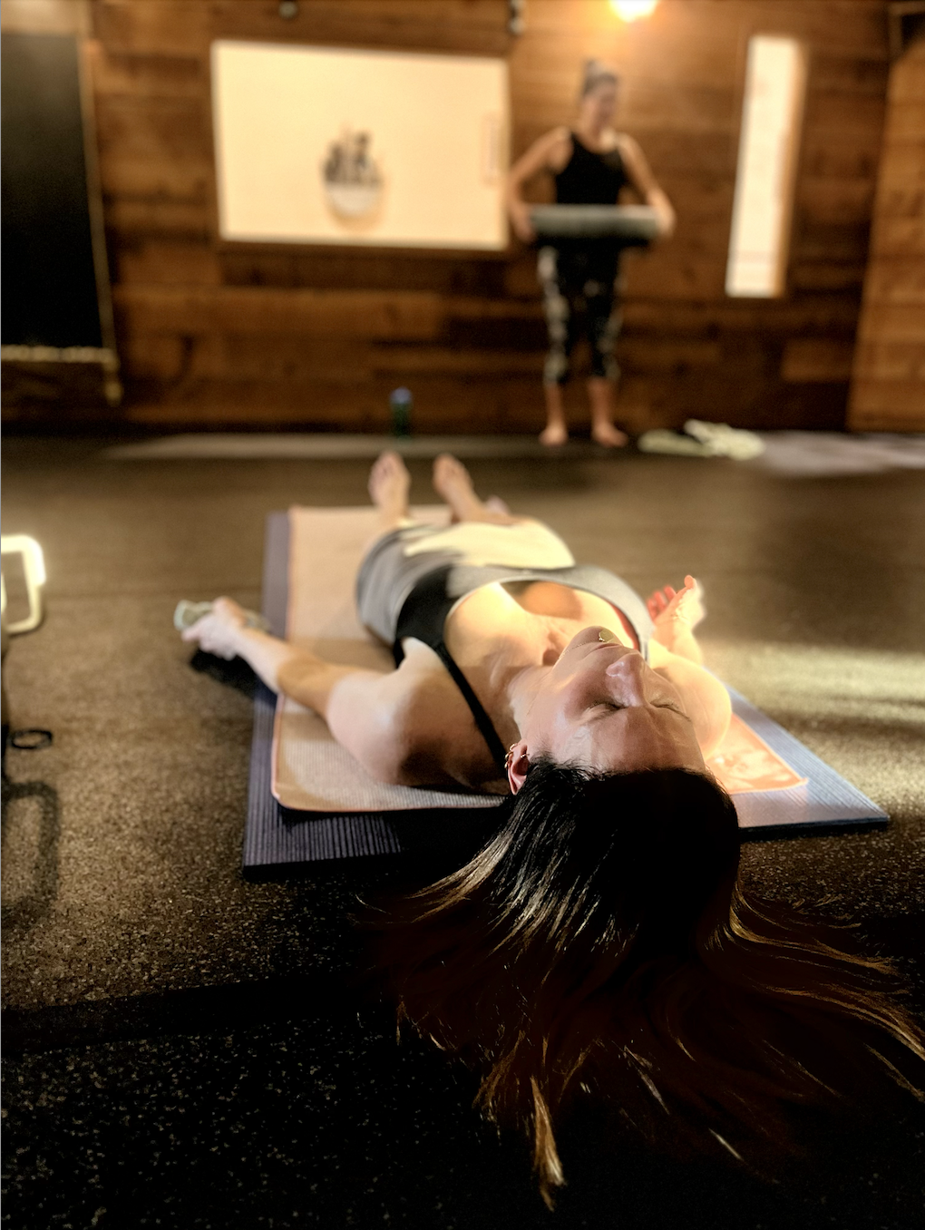 A woman lying on her back on a yoga mat in a yoga class with a woman standing at the front holding a yoga bolster.