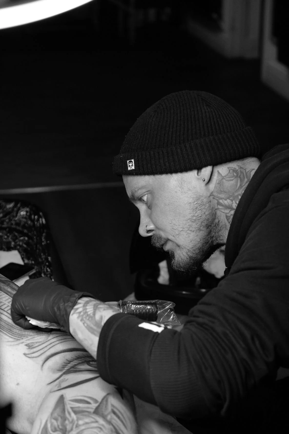 A man with tattoos, a beanie, and a nose piercing is tattooing a person's arm in a tattoo studio.