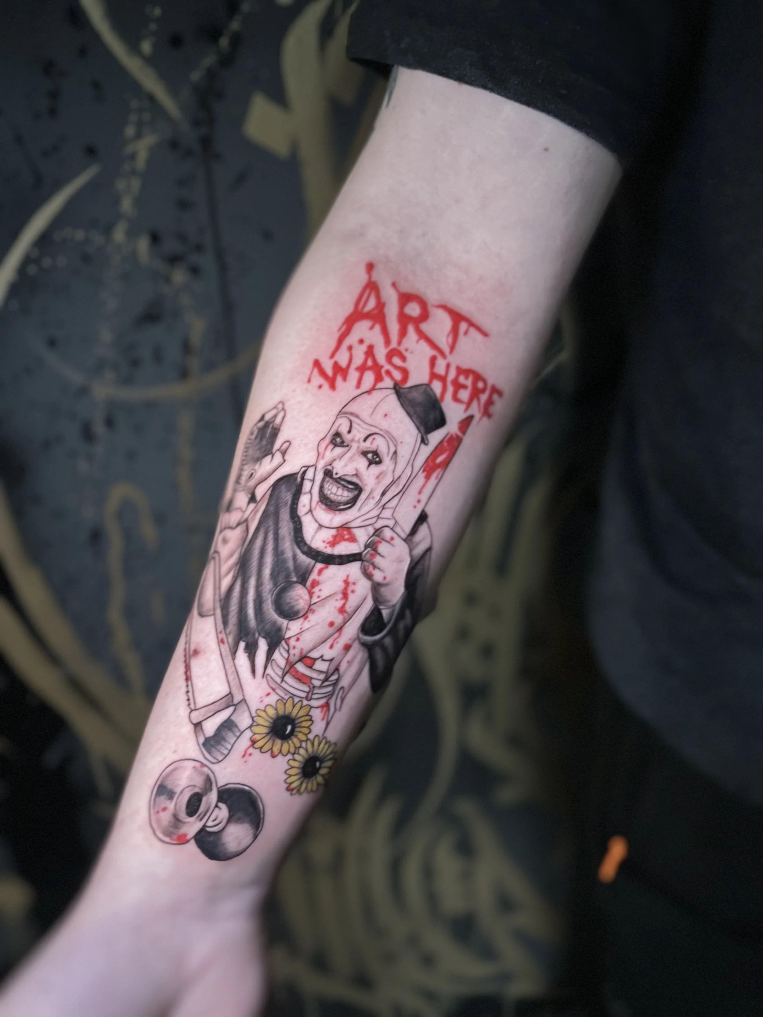 Tattoo of a screaming clown with blood splatter, a skull, and sunflowers, with the phrase "ART WAS HERE" written in red above.