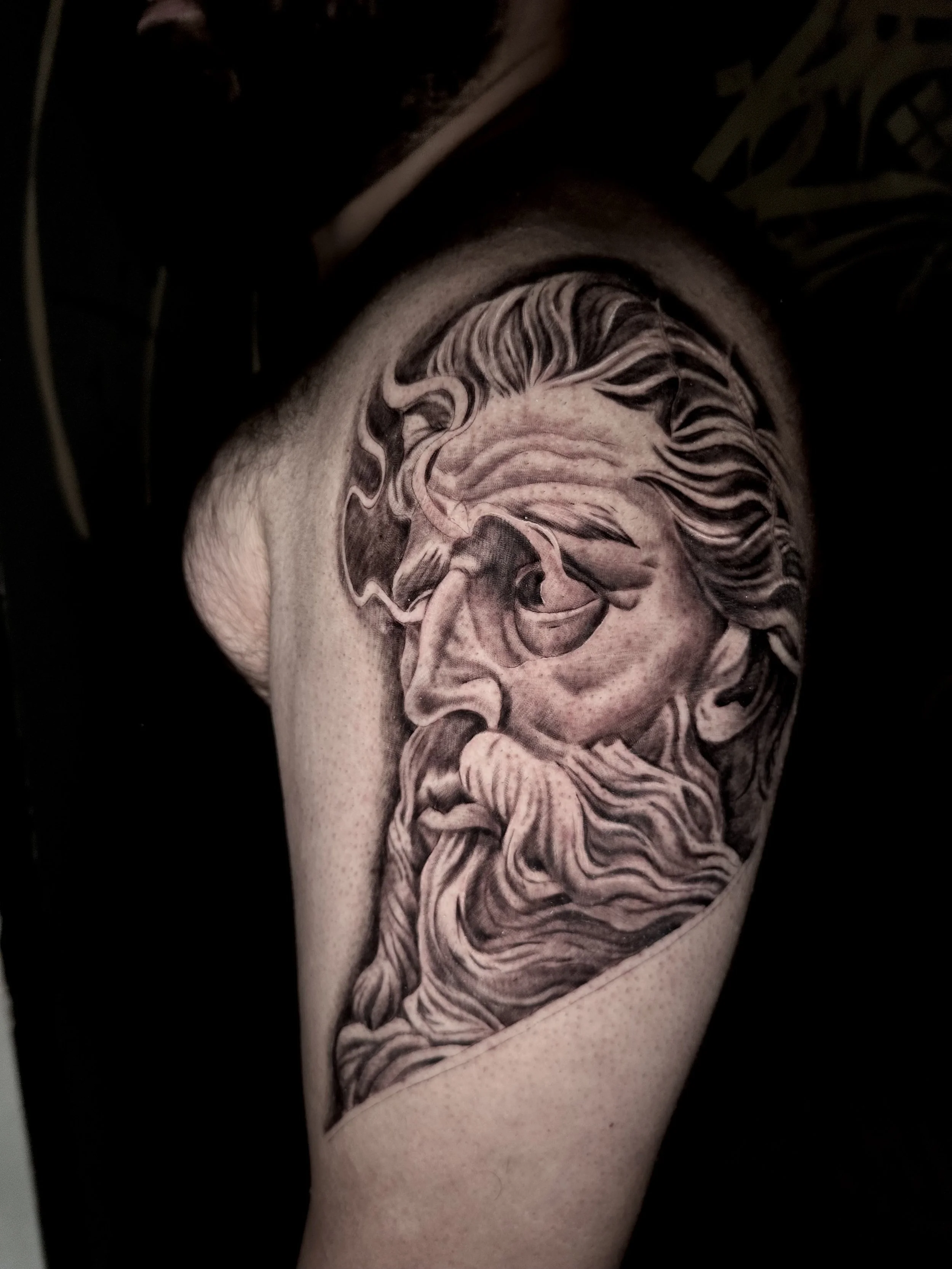 Black and gray tattoo of an ancient Greek or Roman statue's face with a flowing beard and hair, on a person's upper arm.