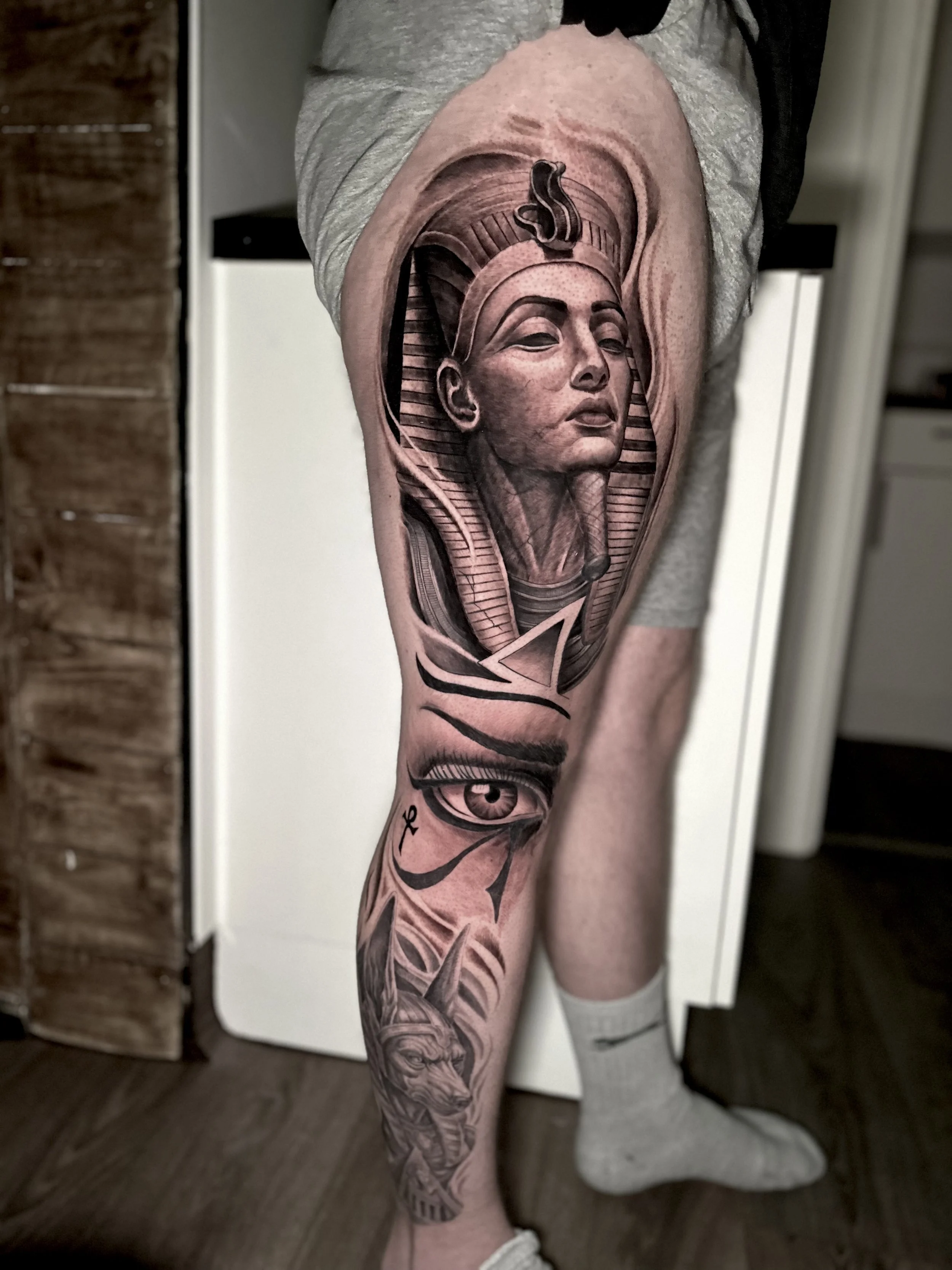 Tattoo of an Egyptian pharaoh's bust, an eye, and a lion's head on a person's leg.