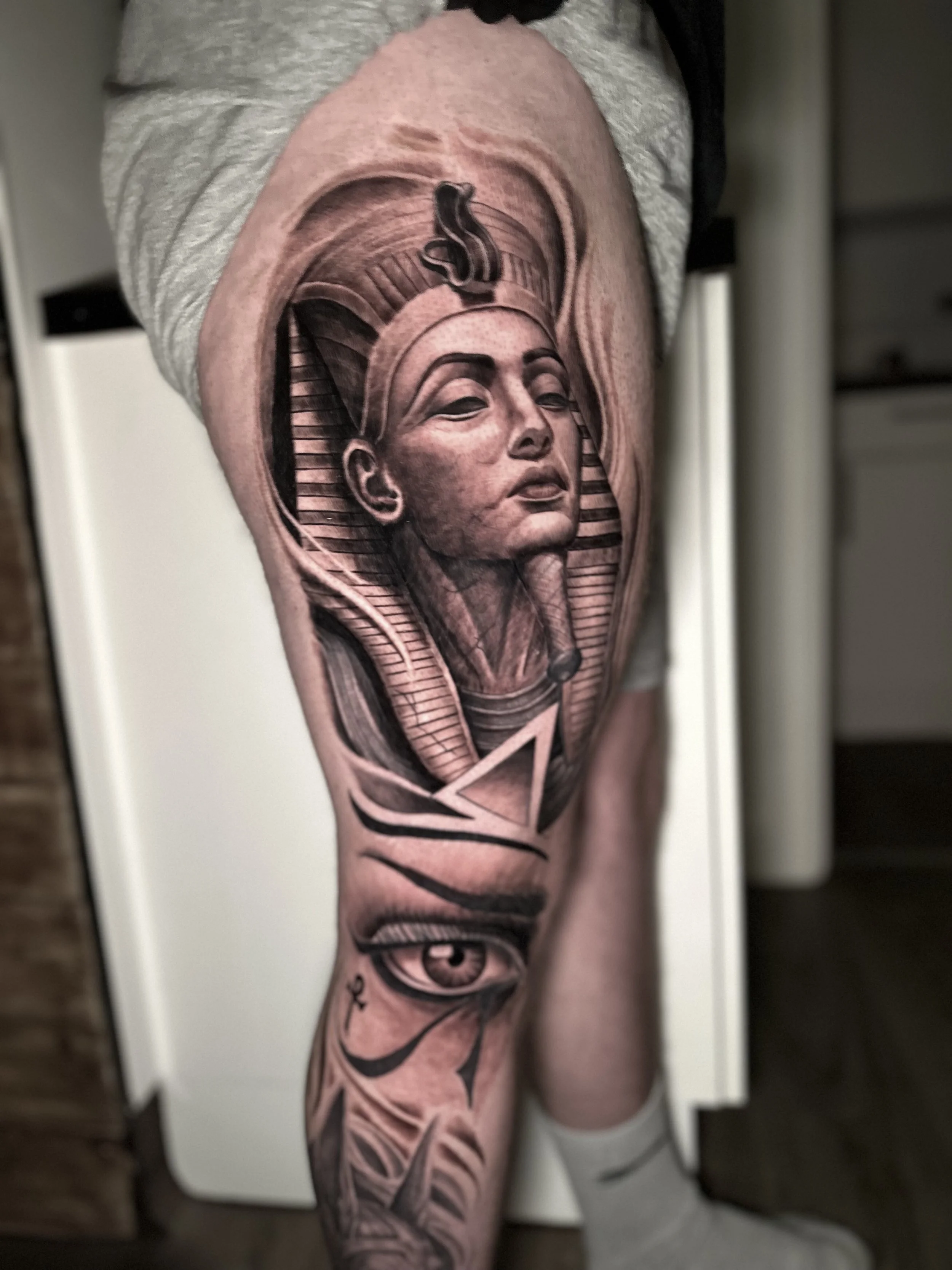 Tattoo of a woman's face wearing a headdress with stairs leading up, under an ornate border. Below, an eye with rays extending outward.