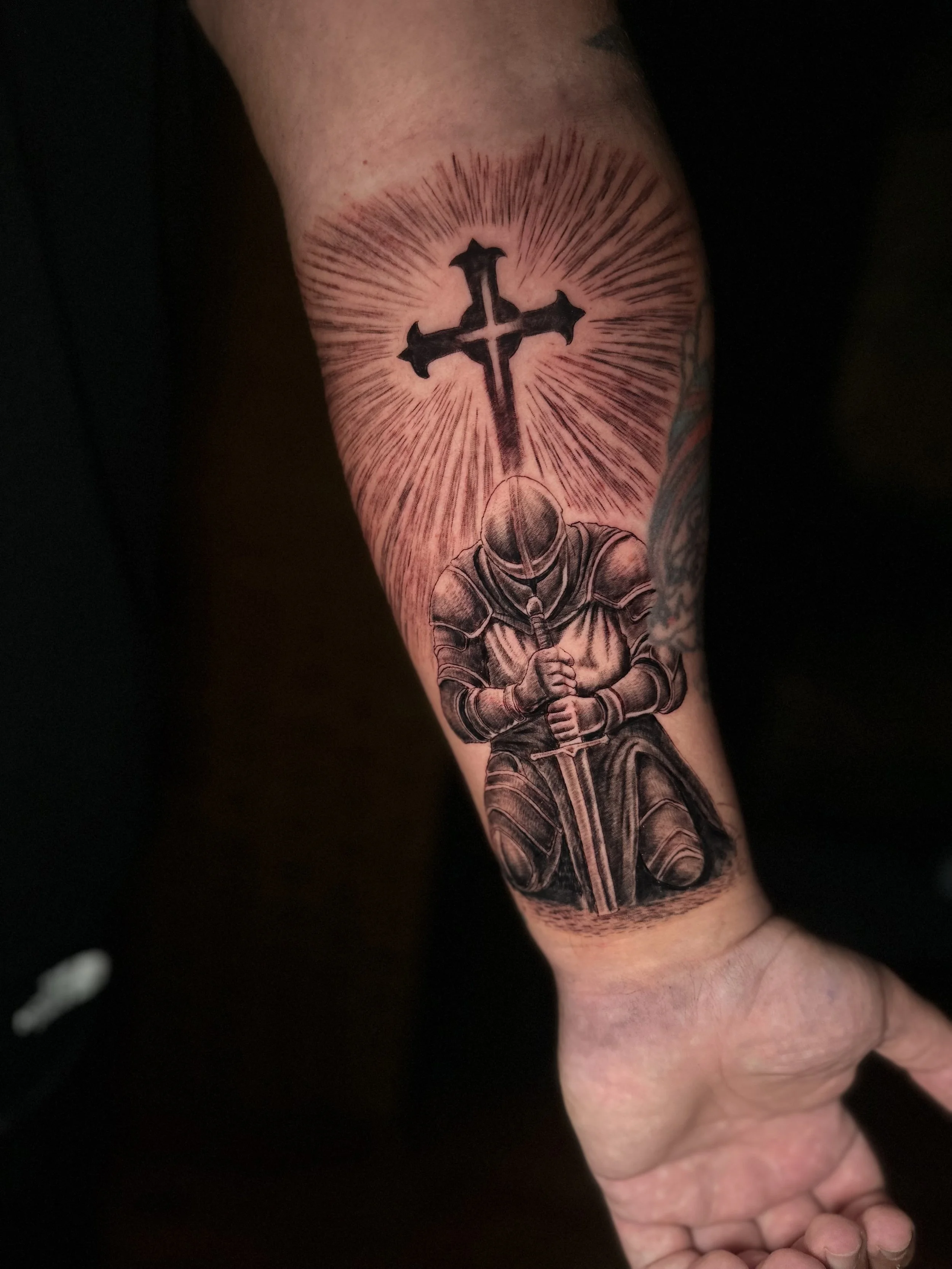 Tattoo of a kneeling knight holding a sword, with a radiant cross above, on a person's forearm.