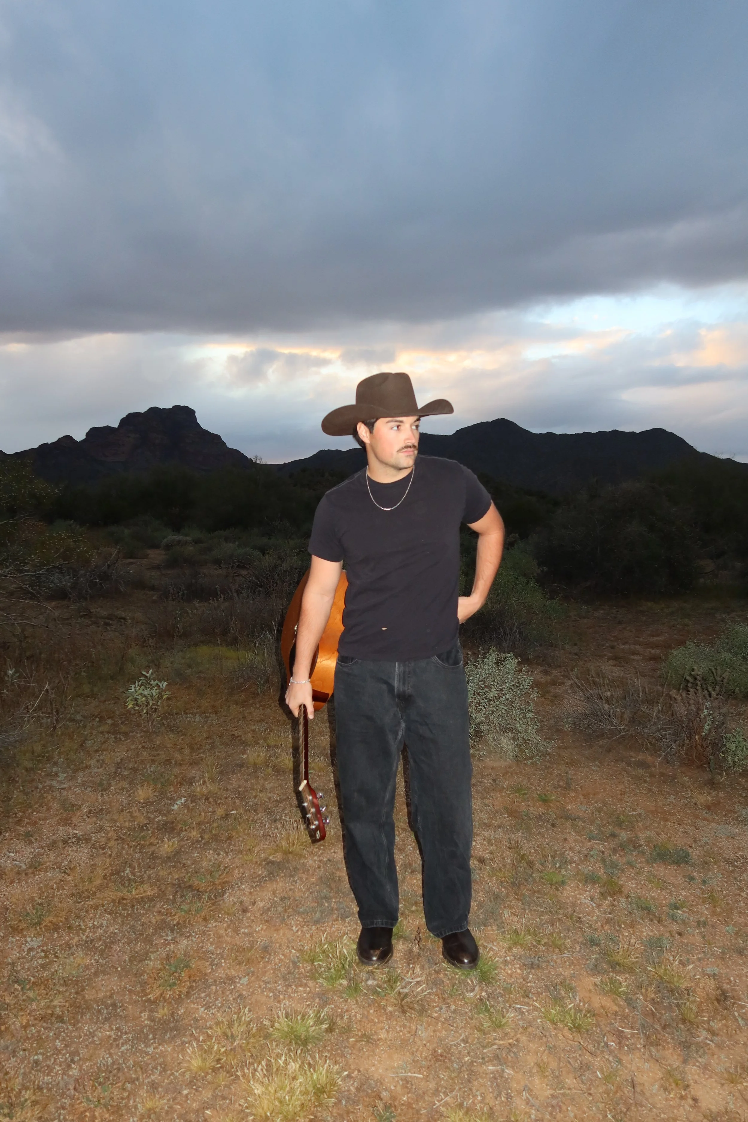 A man wearing a cowboy hat, black t-shirt, and dark jeans standing outdoors on dirt ground with shrubs, holding a guitar, with mountains and cloudy sky in the background.