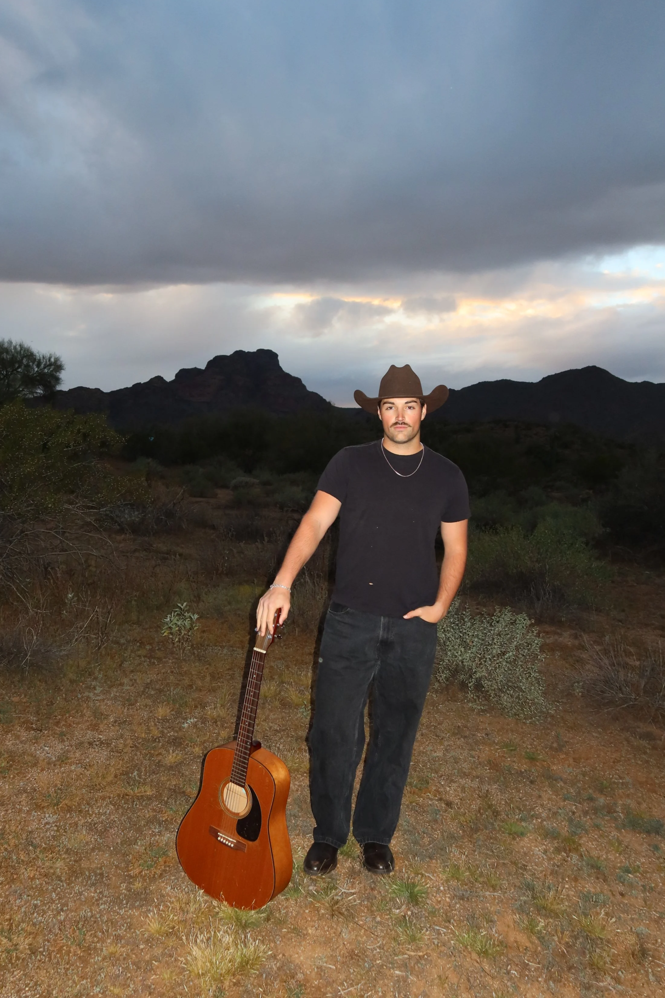 A man in a black t-shirt and jeans standing in a desert landscape with mountains in the background wearing a cowboy hat, holding a guitar, with another guitar on the ground.