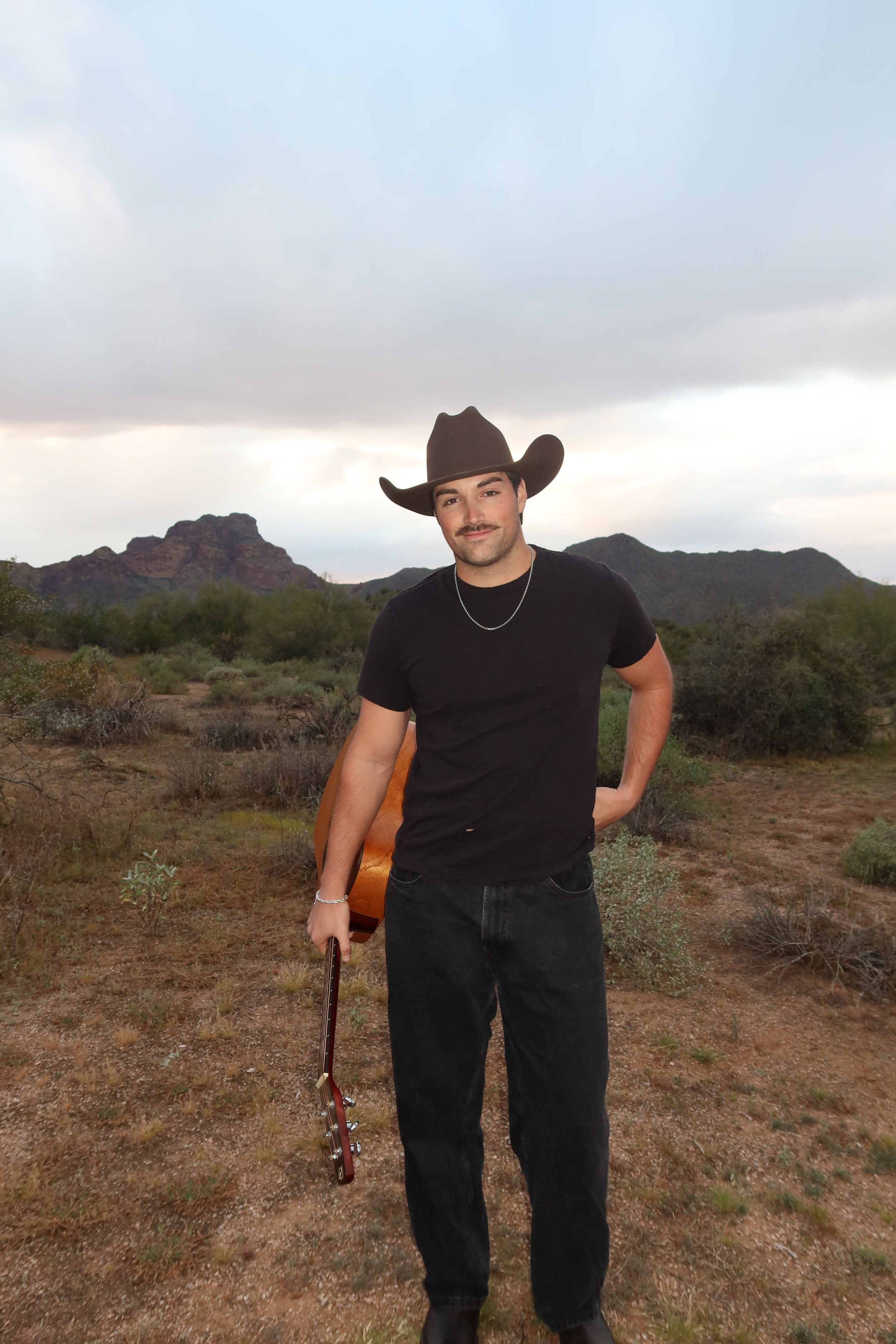 A man with a mustache wearing a cowboy hat, black t-shirt, black pants, and a silver chain, holding a handle of a guitar, standing outdoors in a desert landscape with mountains and overcast sky in the background.