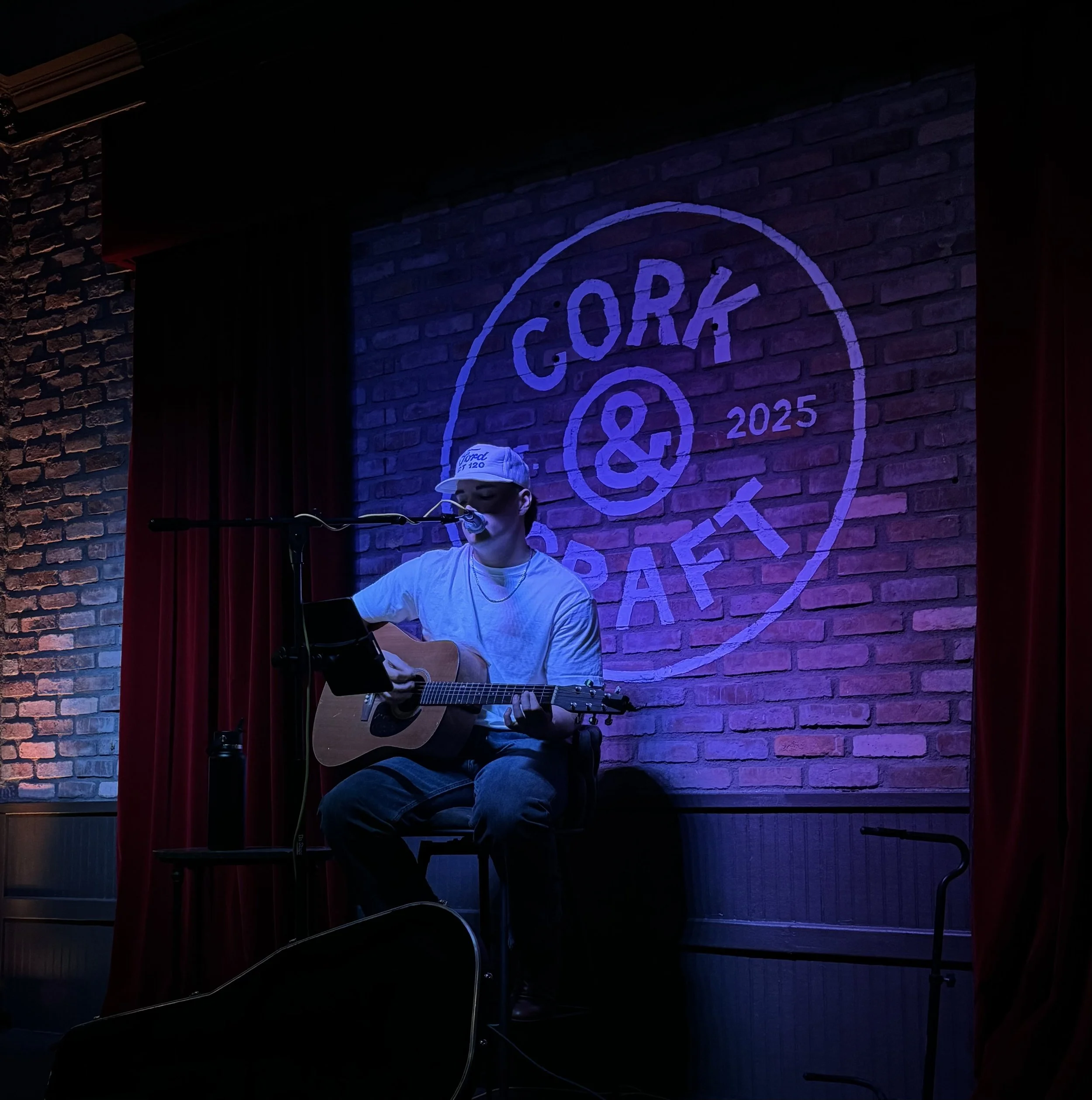 A person wearing a white cap, glasses, and a white shirt plays an acoustic guitar and sings into a microphone on a stage with a brick wall background illuminated with blue and purple lighting, and a logo with the word 'GORE' and the year '2025' behind.