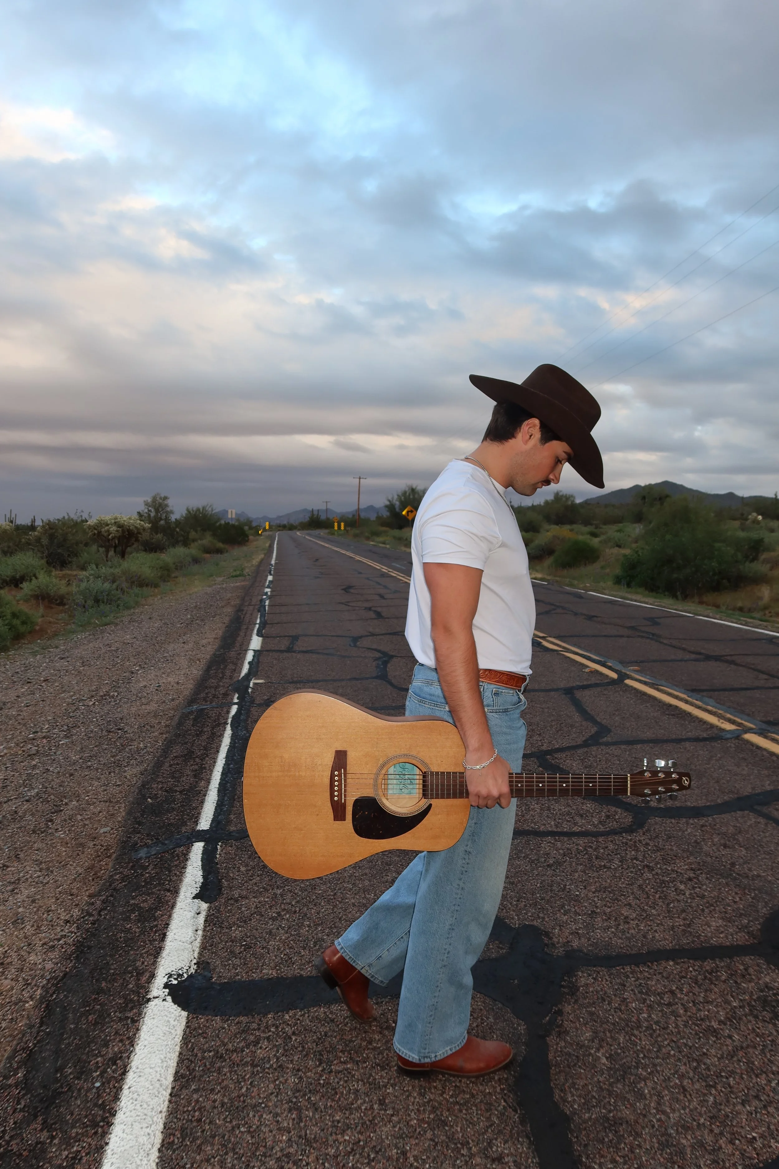 A young man wearing a wide-brimmed cowboy hat, white t-shirt, and jeans, standing on a deserted road with a guitar hanging by his side. The background shows a cloudy sky, desert shrubs, and distant mountains.