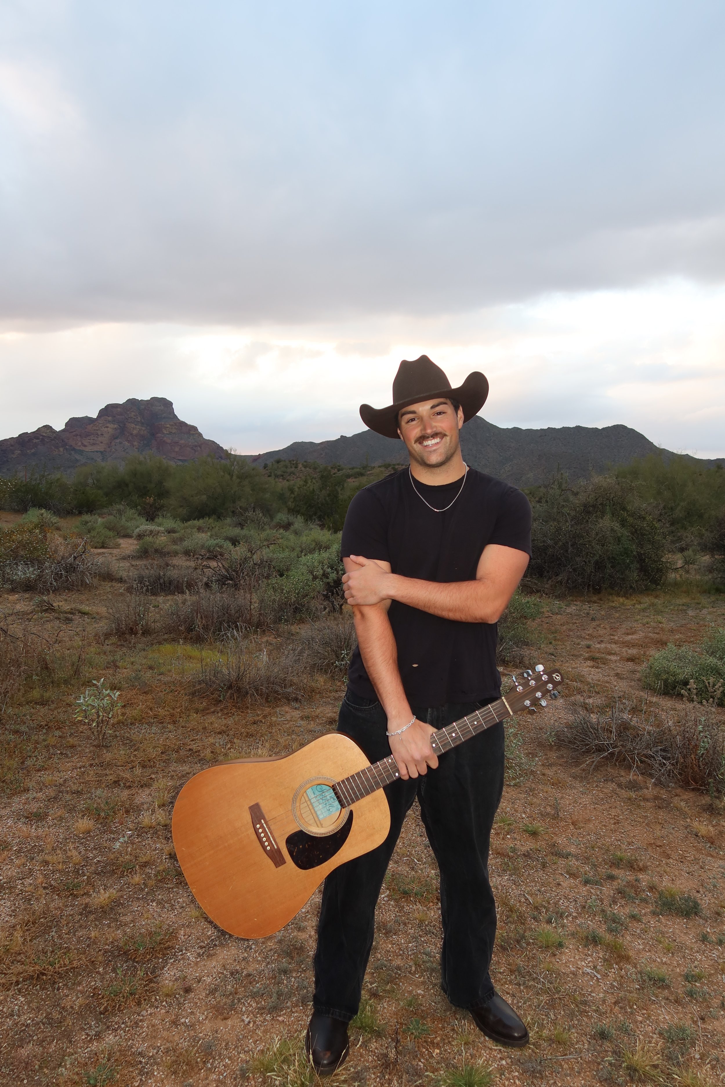 A man wearing a cowboy hat and black clothing, smiling, holding an acoustic guitar in a desert landscape with mountains and shrubs, under a cloudy sky.