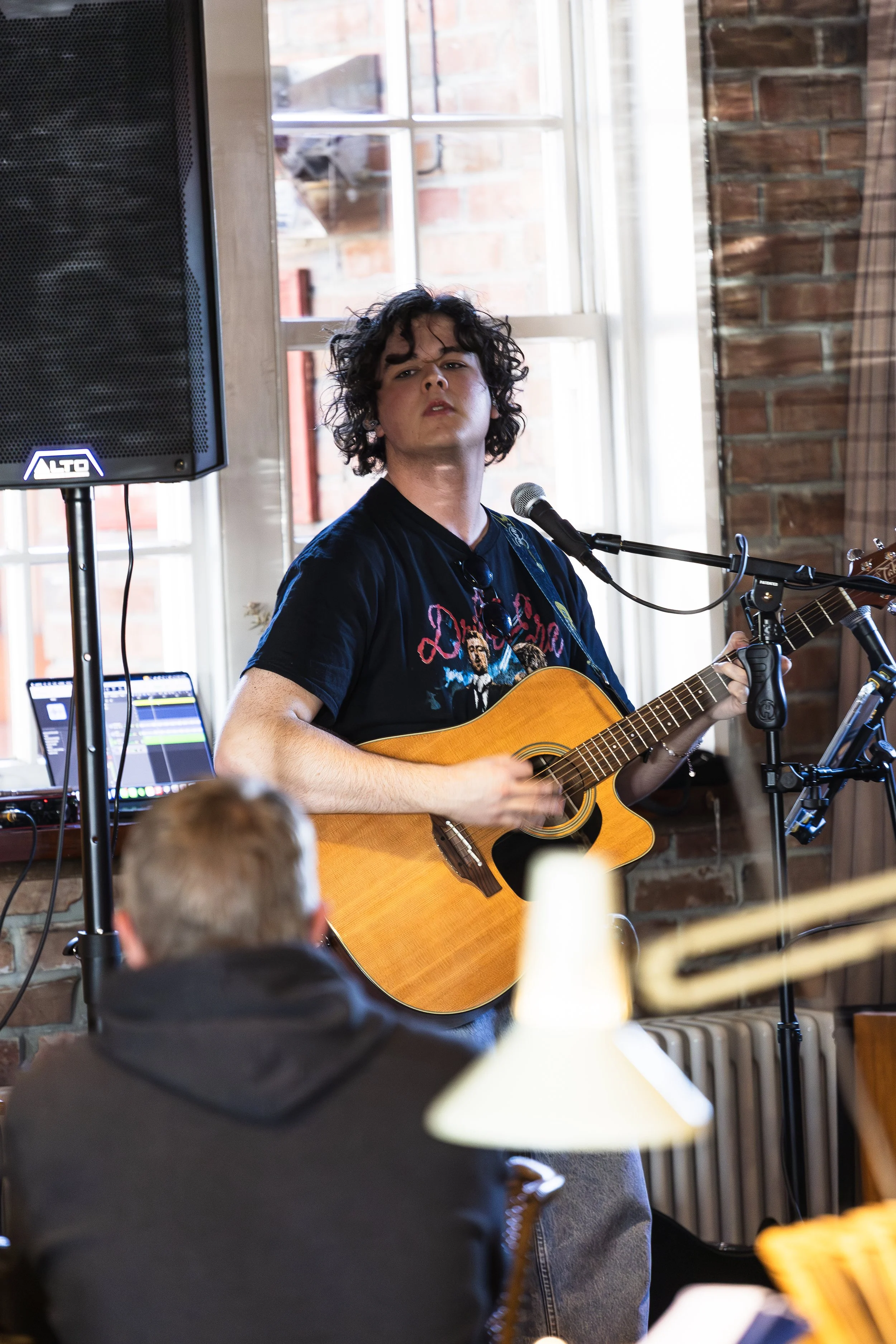 A young man with curly dark hair performing live music indoors with a guitar, microphone, and audio equipment, in front of a brick wall and a large window.