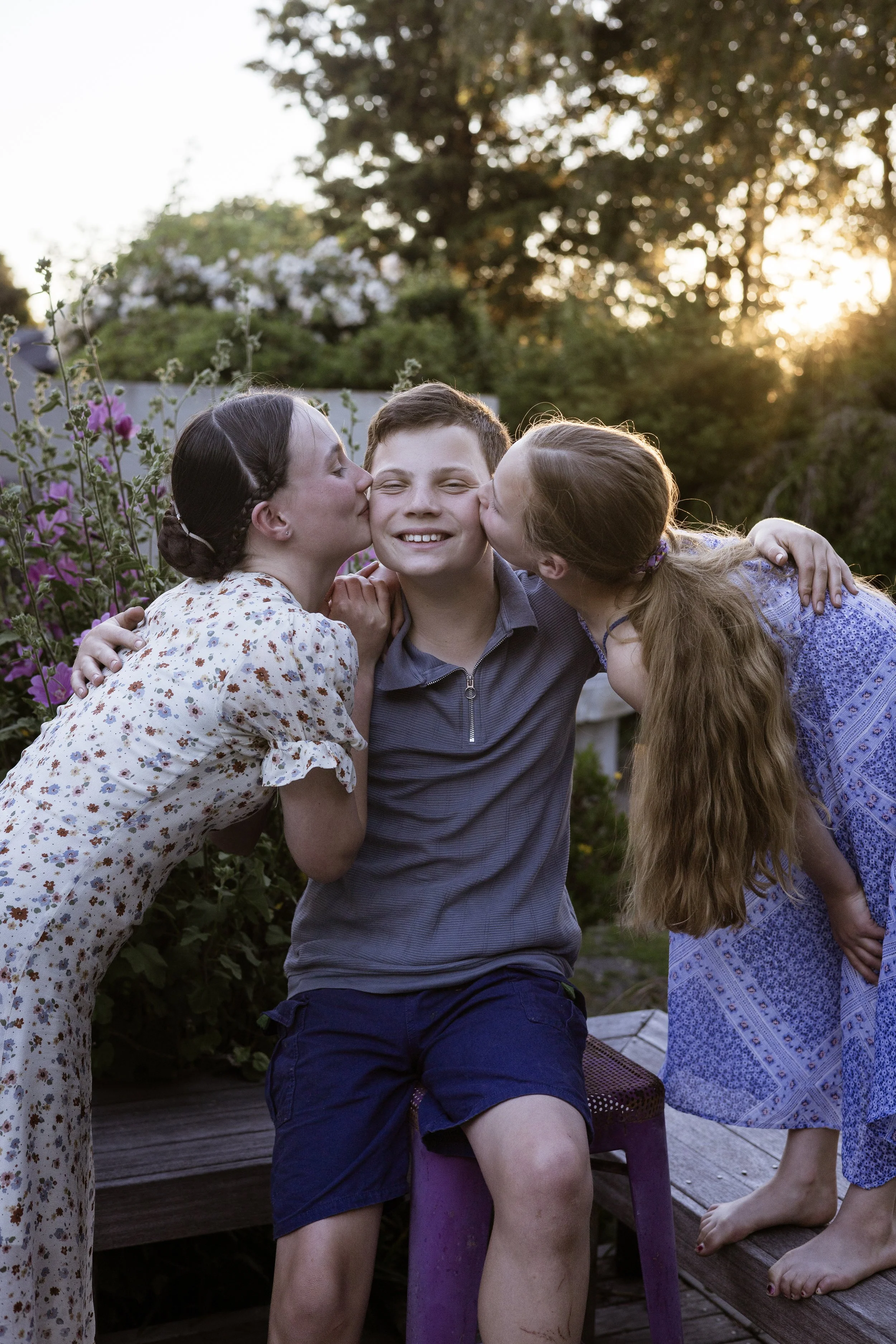 Three children, two girls and one boy, outdoors during sunset. The girls are kissing the boy on the cheeks. The boy has a big smile. The setting features trees, flowers, and a wooden deck.