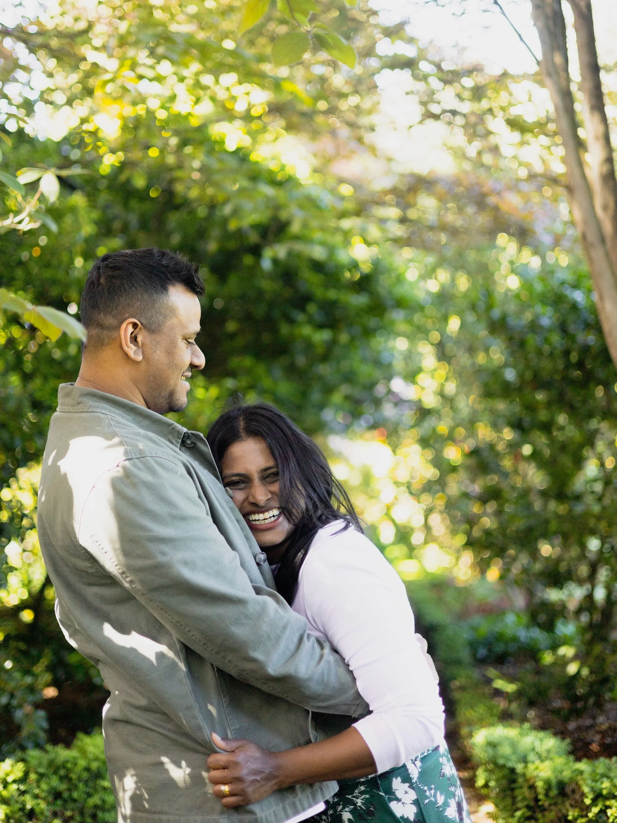 A happy couple hugging and smiling outdoors with lush green trees and sunlight in the background.