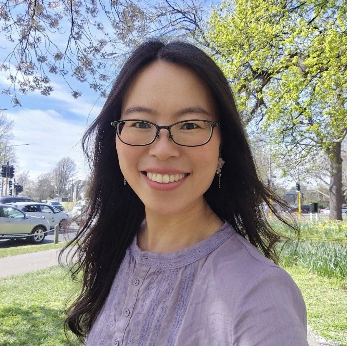 Smiling woman with glasses and earrings outdoors on a sunny day, with trees, cars, and a traffic light in the background.