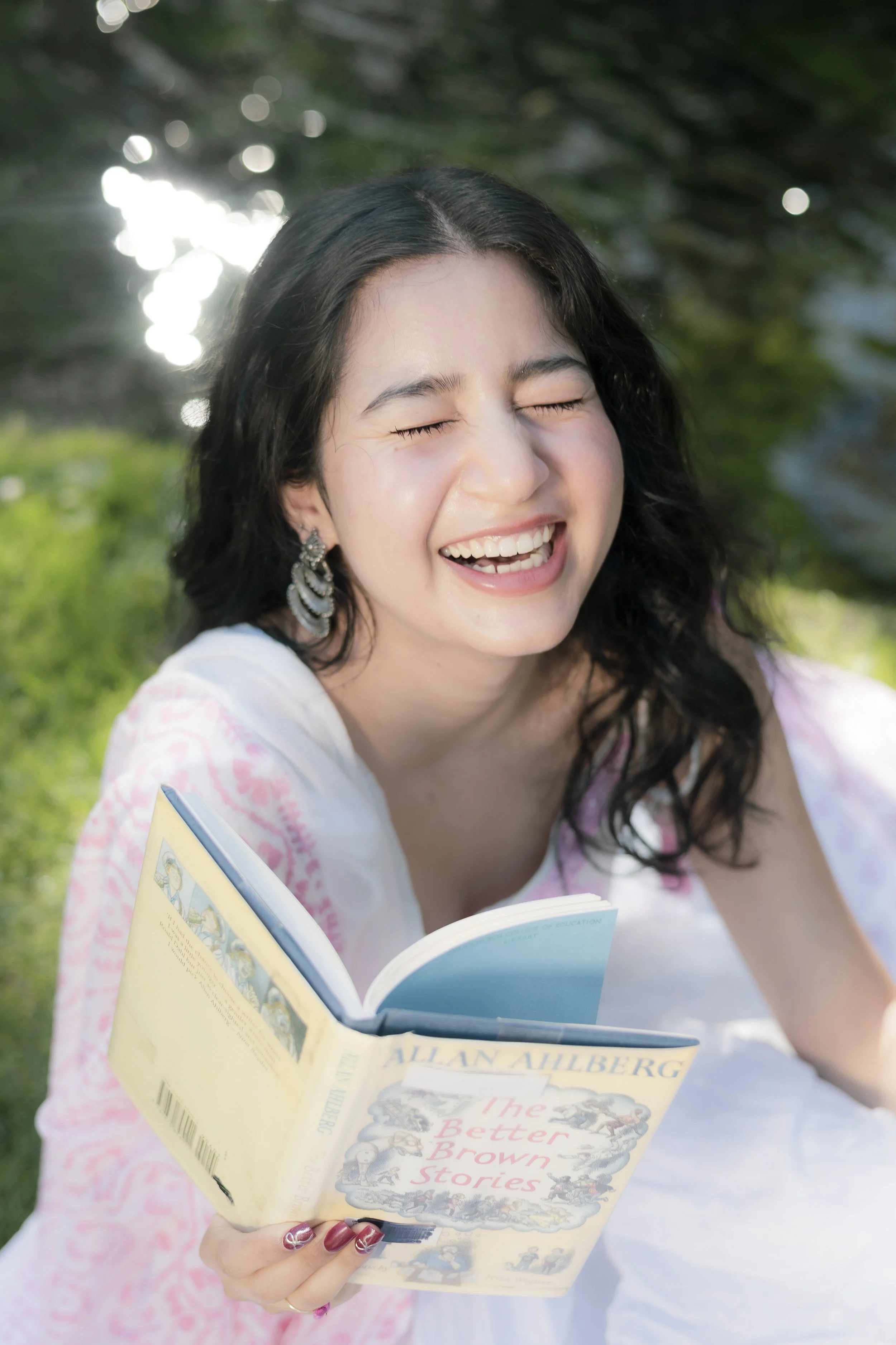 A young woman with dark, wavy hair and large earrings is smiling with her eyes closed, holding an open book titled "The Better Brown Stories" by Allan Ahlberg, outdoors near a body of water with trees in the background.