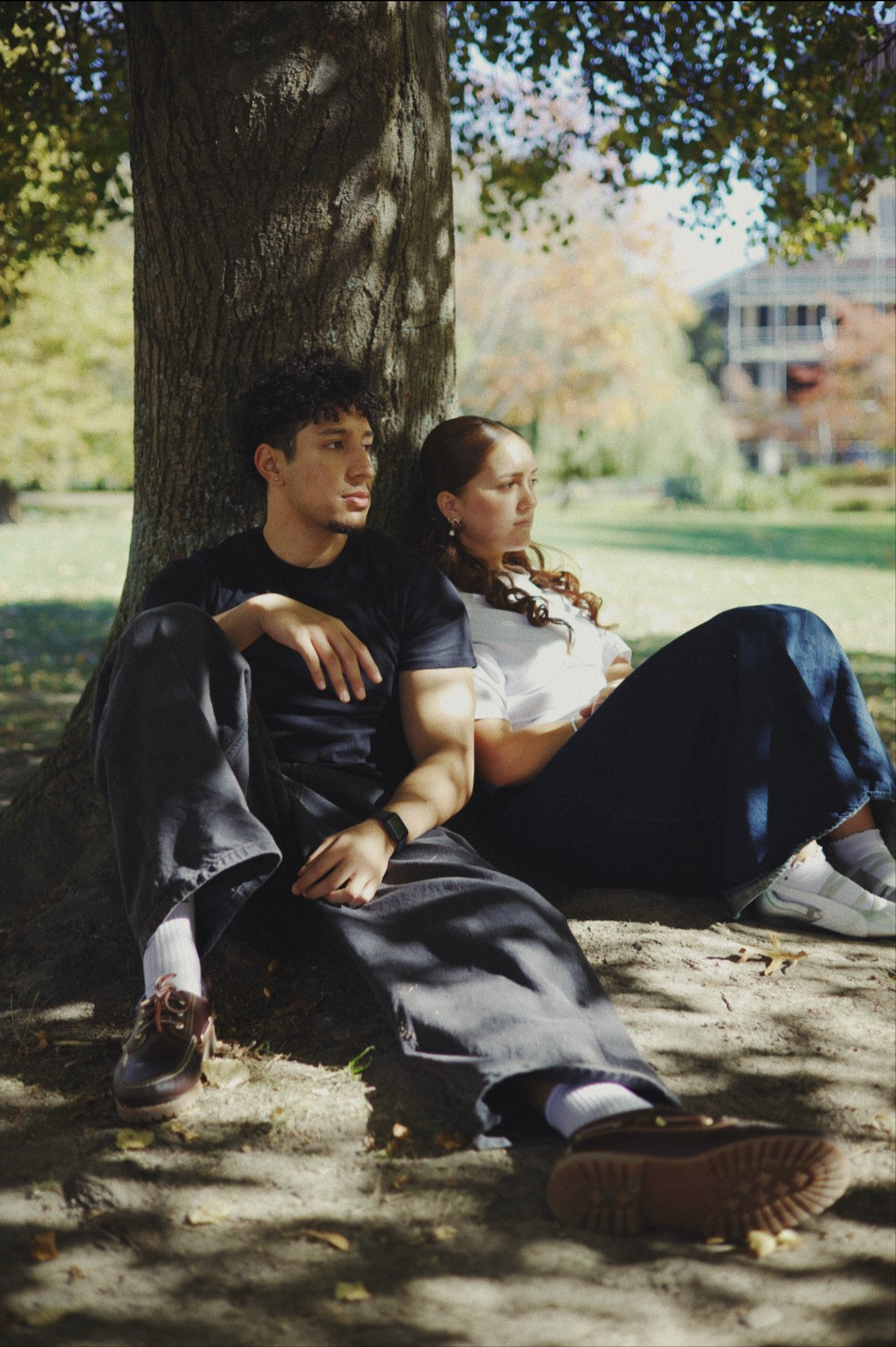 A young man and woman sitting on the ground under a tree in a park, leaning against the trunk of the tree with leaves and grass in the background, on a sunny day.