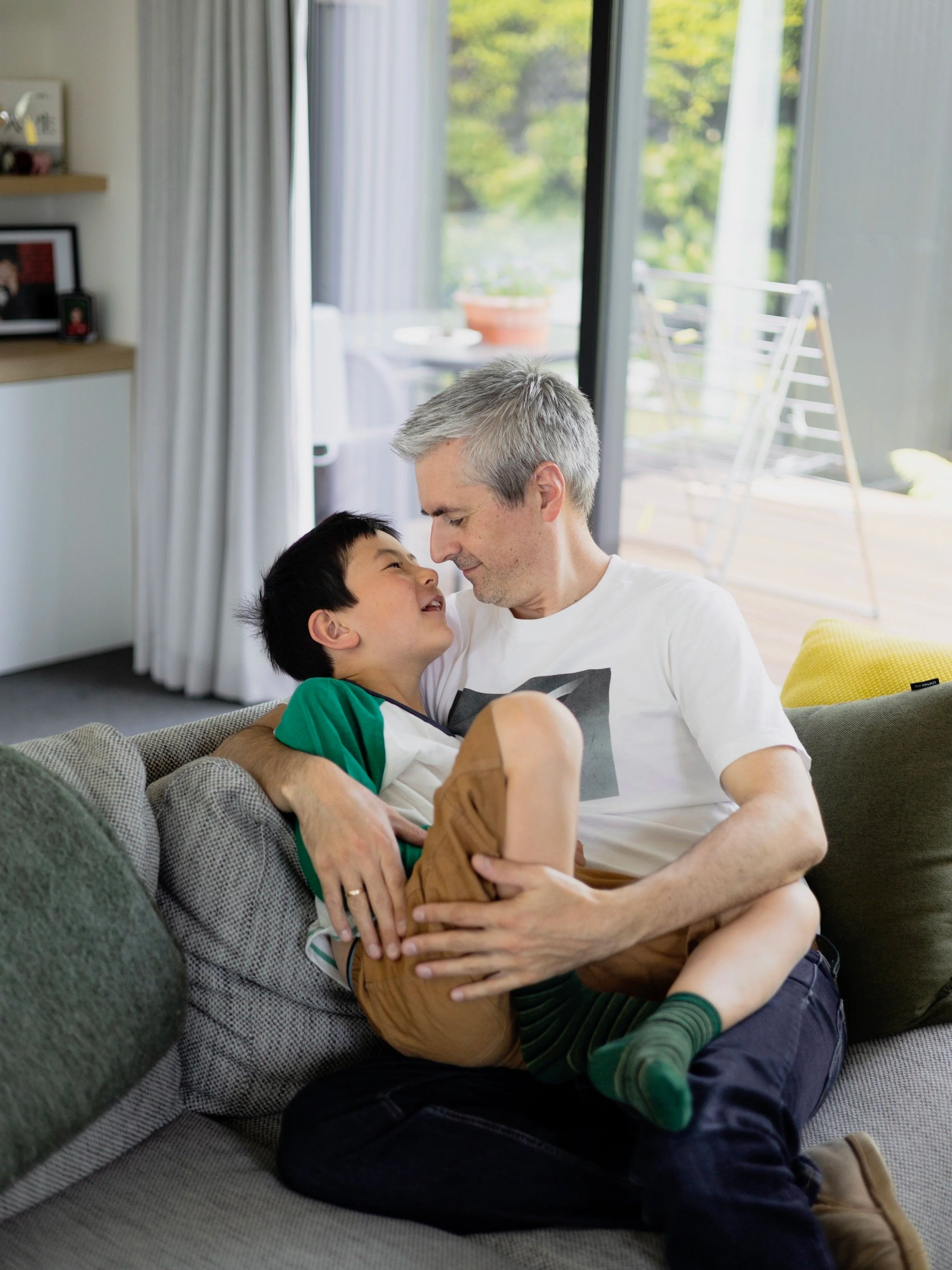 A man and a boy sitting on a sofa, smiling and touching foreheads; the man is holding the boy in his arms indoors near large sliding glass doors with a view of a balcony.