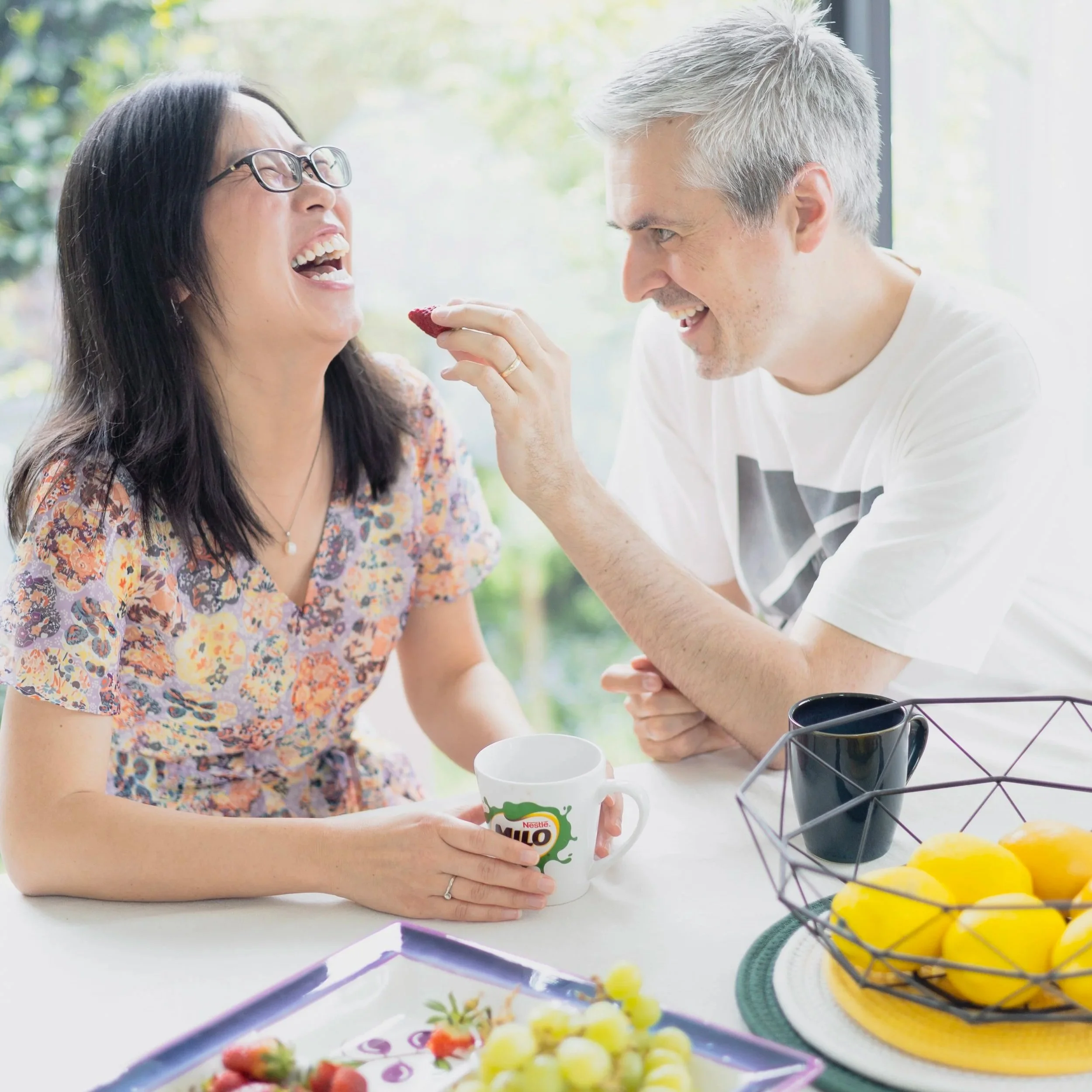 A man and woman laughing and sharing a moment at a breakfast table with fruit and a mug.