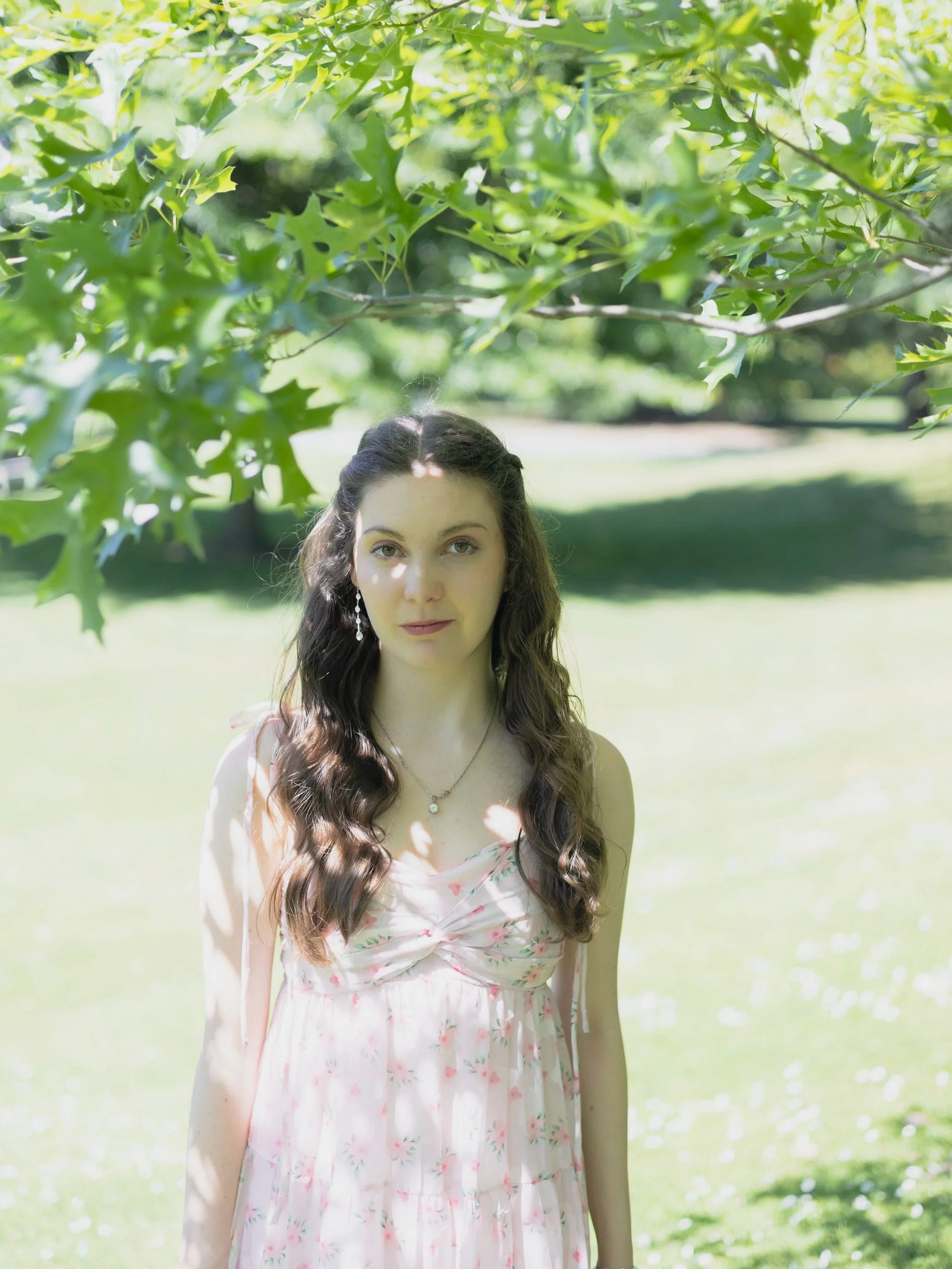 A young woman with long brown hair and light skin standing outdoors in a park, partially shaded by green leaves overhead. She is wearing a light pink floral dress and jewelry.