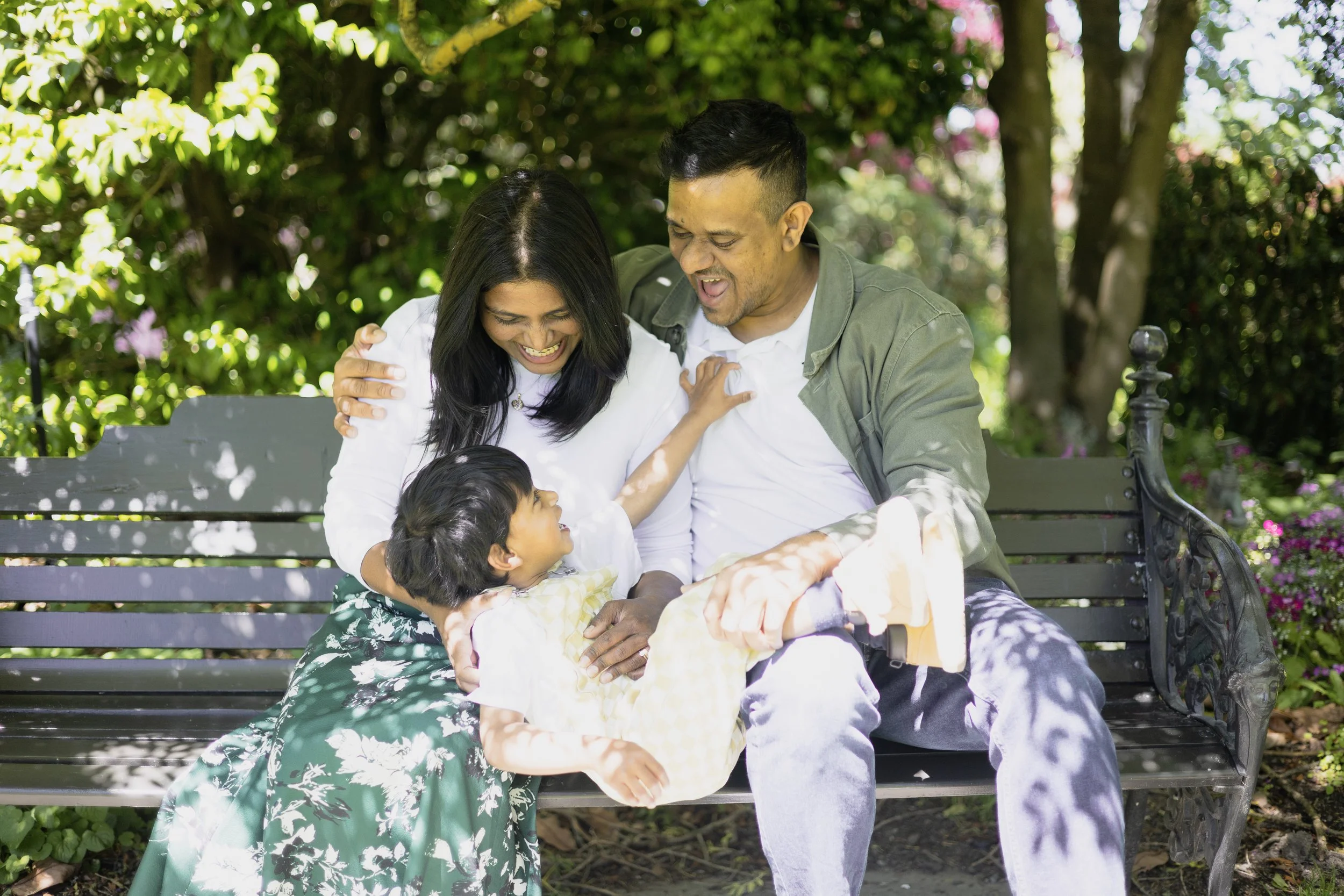 A family of four is sitting on a park bench, laughing and enjoying each other's company. The scene is surrounded by lush greenery and trees, with sunlight filtering through the leaves.
