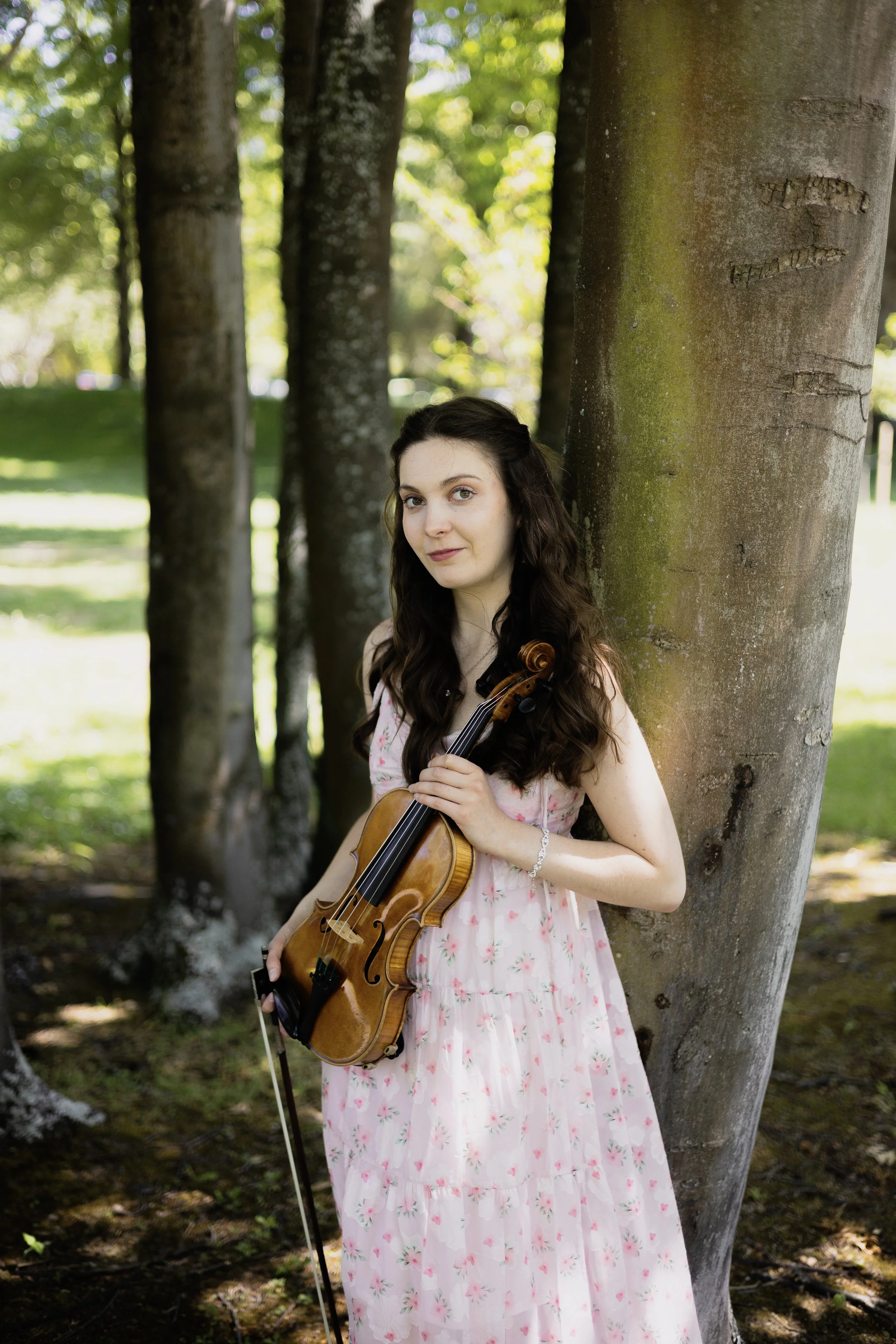 A young woman with long dark curly hair, wearing a pink floral dress, standing outdoors by a large tree trunk, holding a violin in her hands, with green trees and grass in the background.