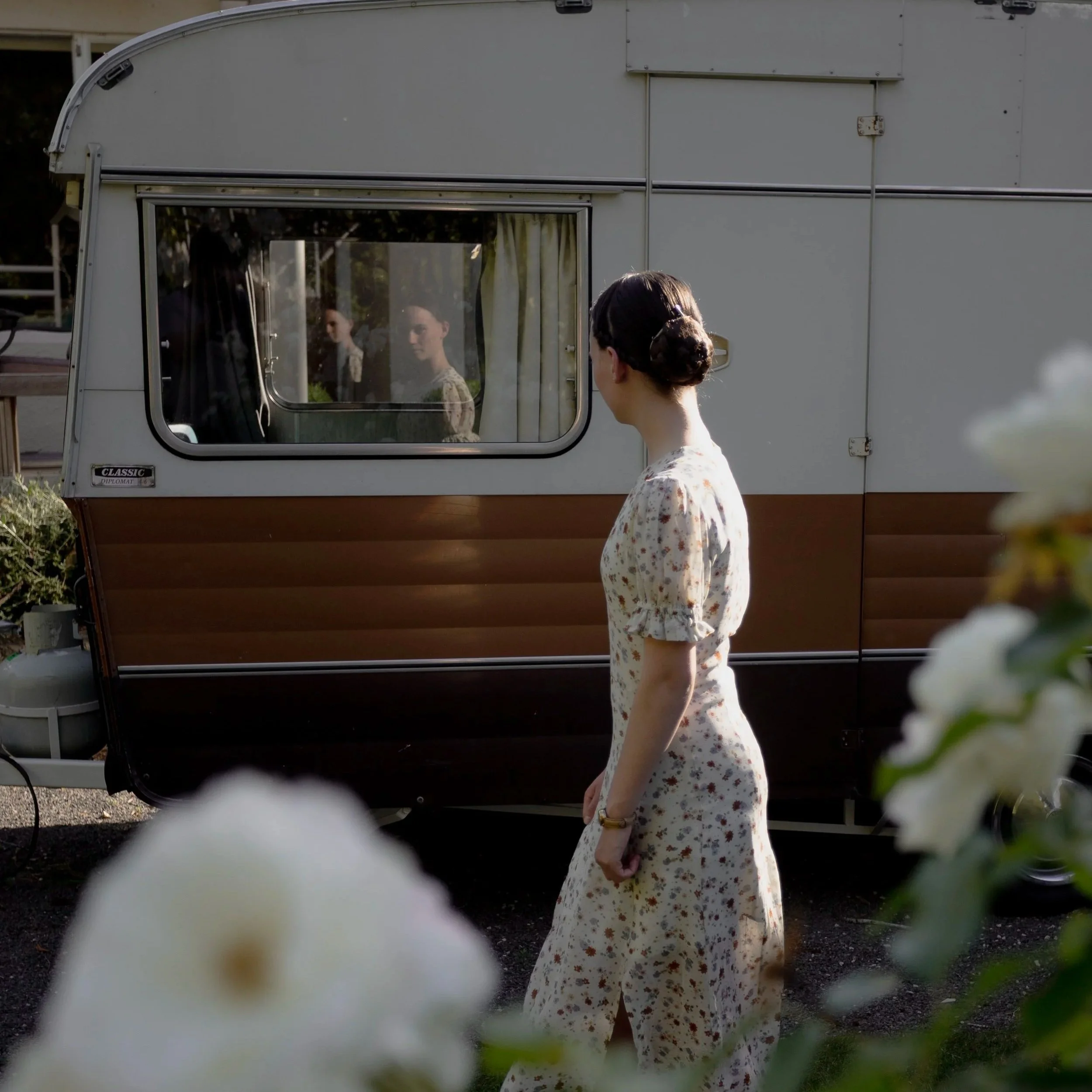 A woman in a floral dress standing outside near a vintage camper trailer, looking at two women inside the trailer through the window, with blurred white flowers in the foreground.