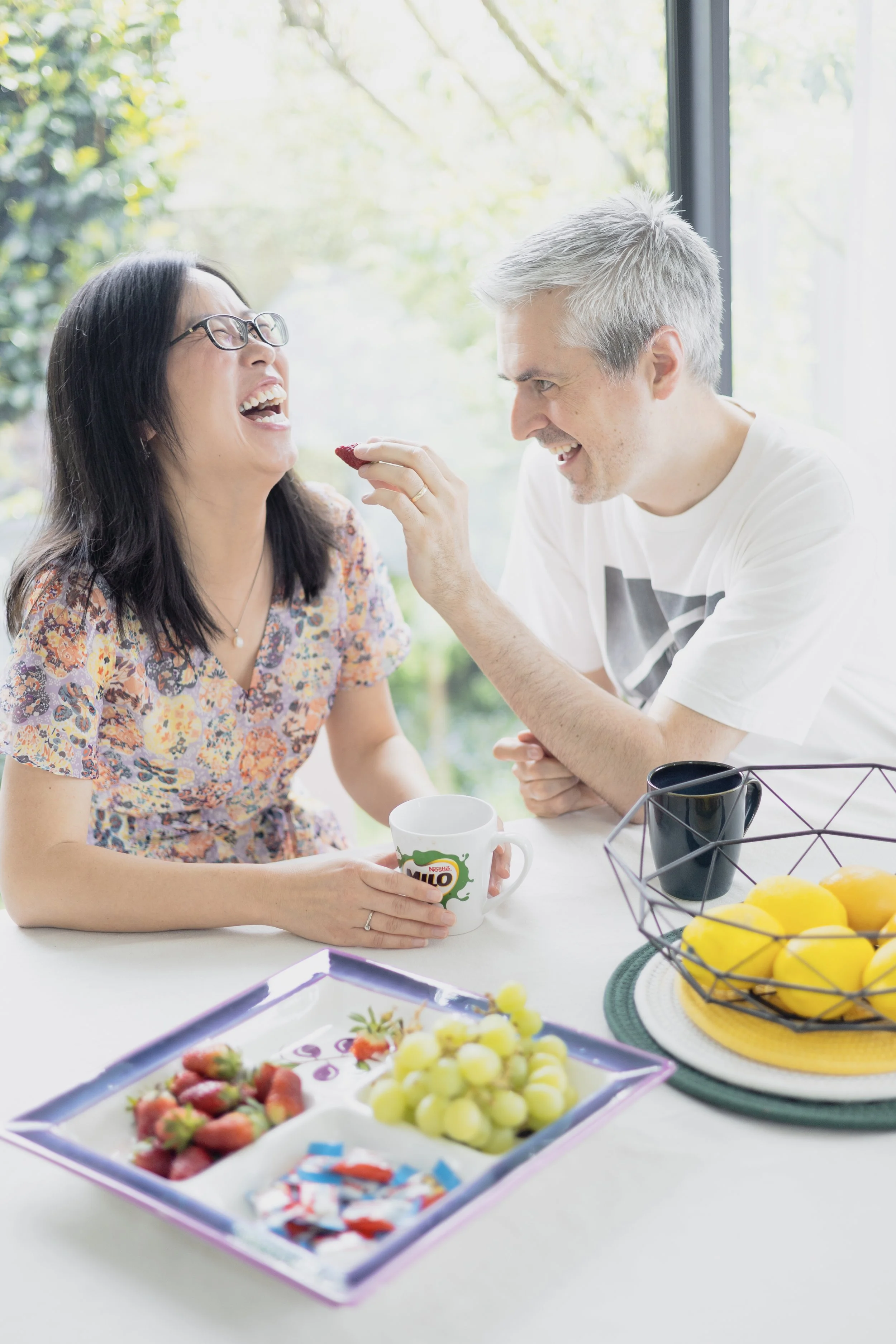 A man feeds a woman with a strawberry while they both sit at a table with bowls of fruit, mugs, and a tray of snacks in a bright room with large windows and greenery outside.