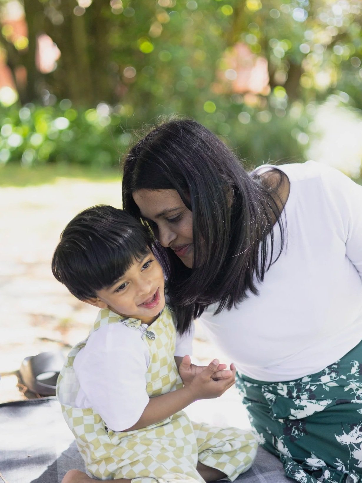 A woman and a young boy sharing a tender moment outdoors, smiling and leaning on each other with a background of greenery.