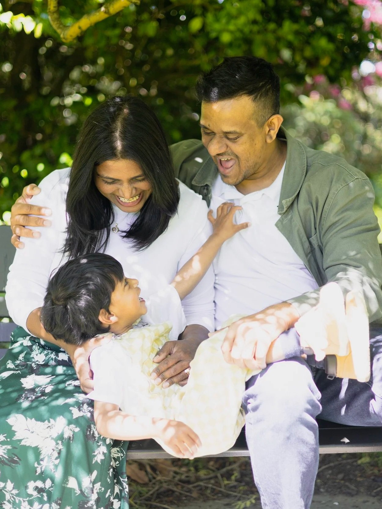 A happy family of three, including a woman, a man, and a young girl, enjoying a moment outdoors on a park bench with trees and greenery in the background.
