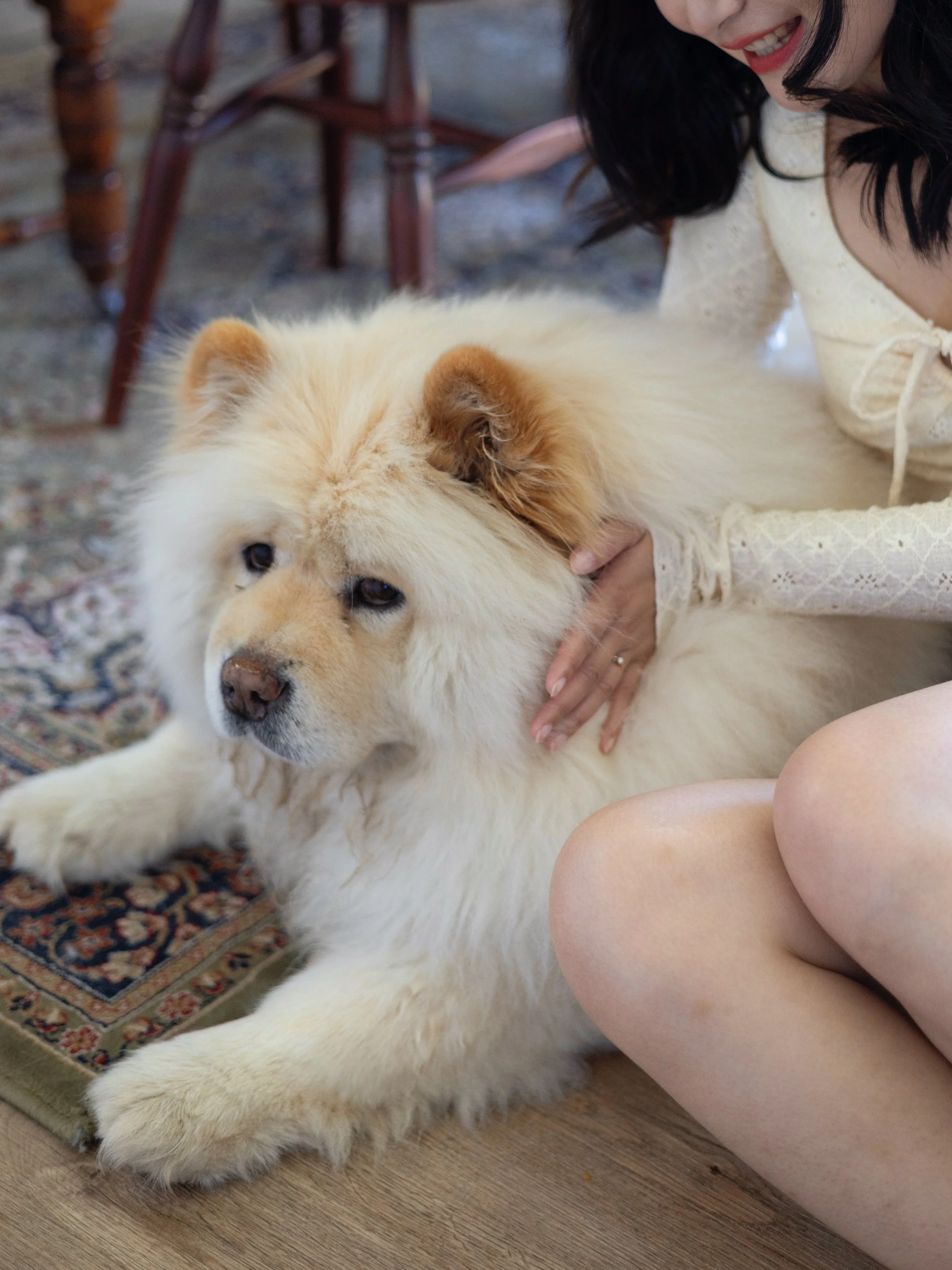 A woman with black hair and a light-colored dress petting a large, fluffy, cream-colored dog on a patterned rug.