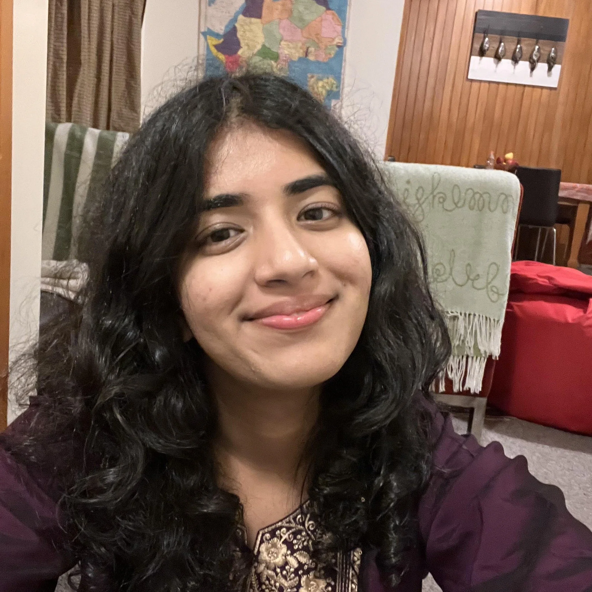 A woman with dark, curly hair smiling in a room with a map of Africa and a wooden panel wall behind her.