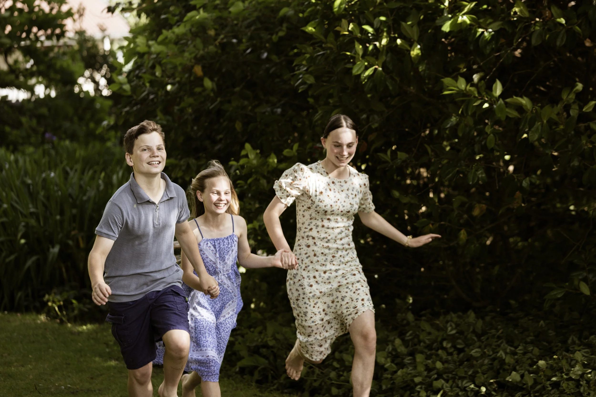 Three children, a boy and two girls, are running outdoors on green grass with a dense bushy background, all smiling and holding hands.
