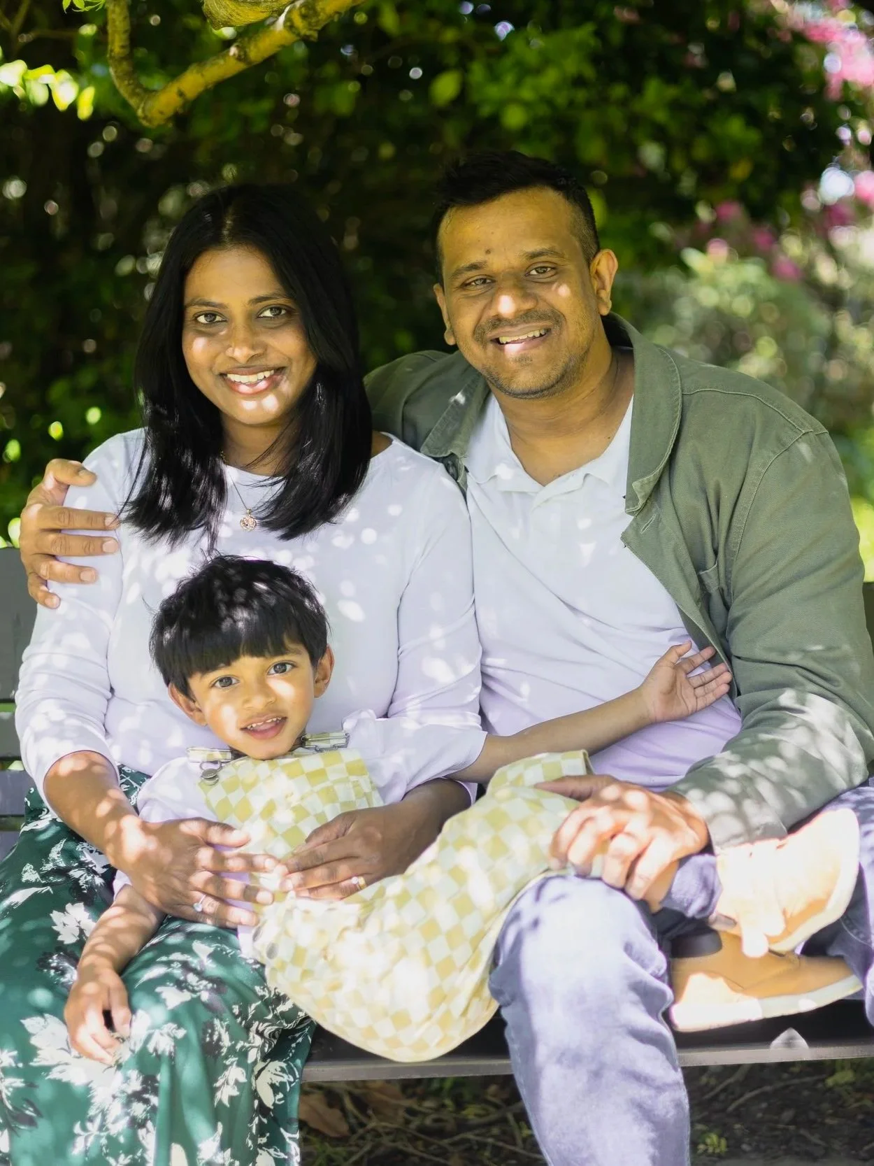 A family of three sitting on a bench outdoors, smiling, surrounded by greenery and pink flowers.