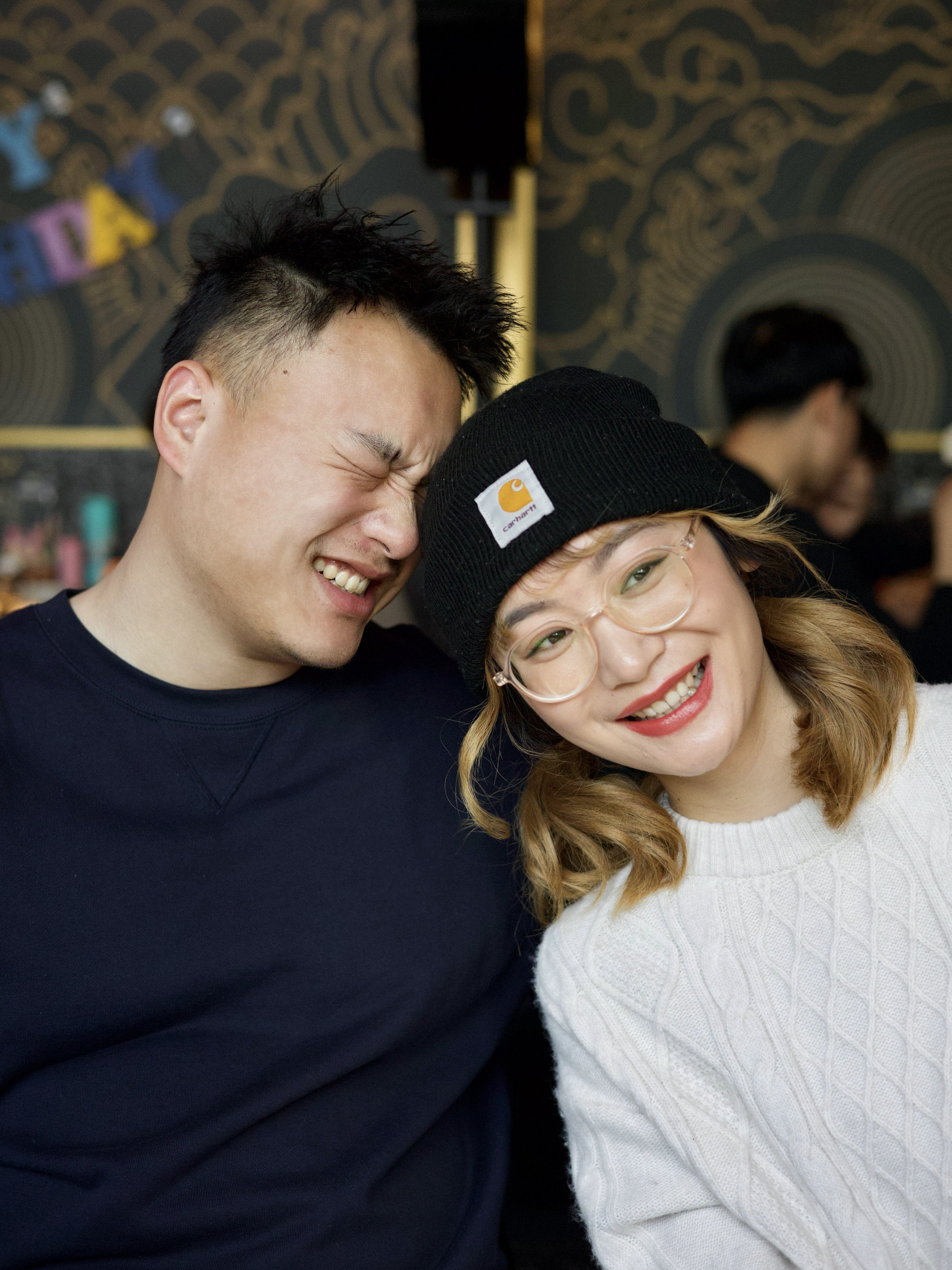 Two friends laughing affectionately, with the man closing his eyes and the woman smiling, in a cozy indoor setting.