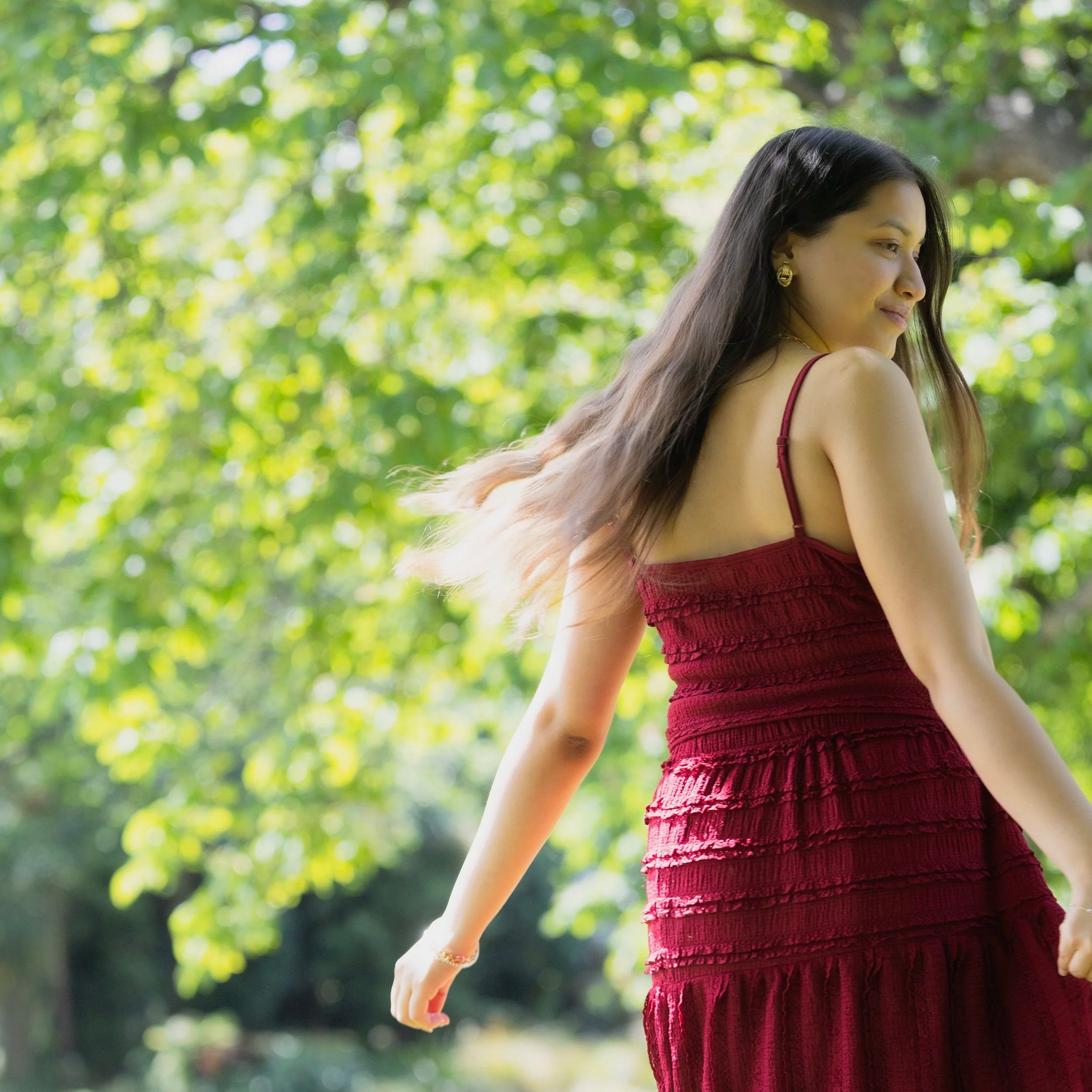 A woman dressed in a red textured swimsuit with long dark hair and gold earrings is standing outdoors in a lush green environment with sunlight filtering through trees.