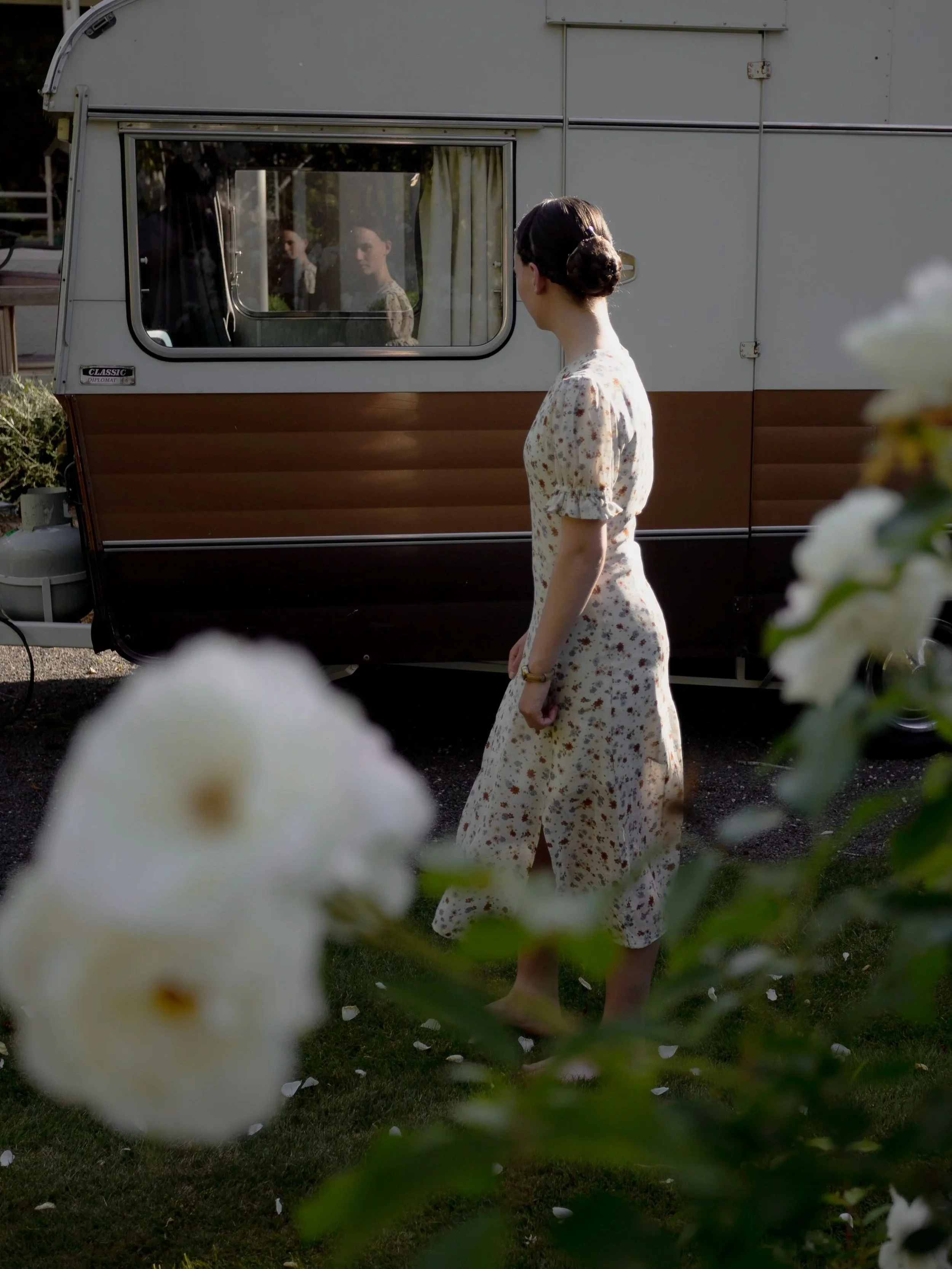A woman with dark hair in a bun wearing a white floral dress stands outside near a caravan, looking at her reflection in the window.