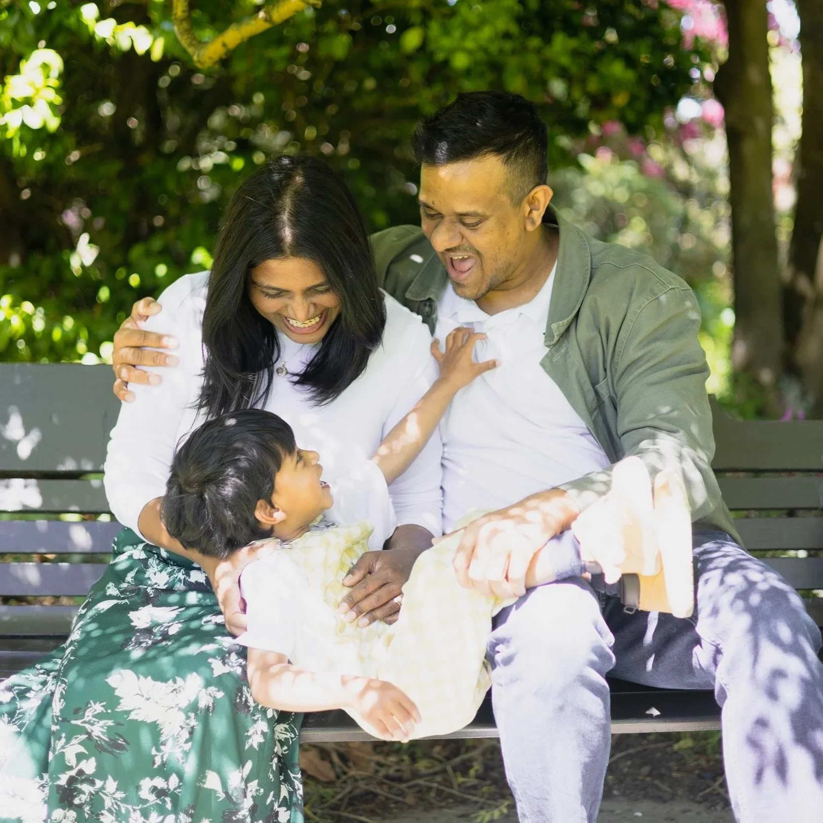 A family of three sitting on a bench outdoors, smiling, surrounded by greenery and pink flowers.
