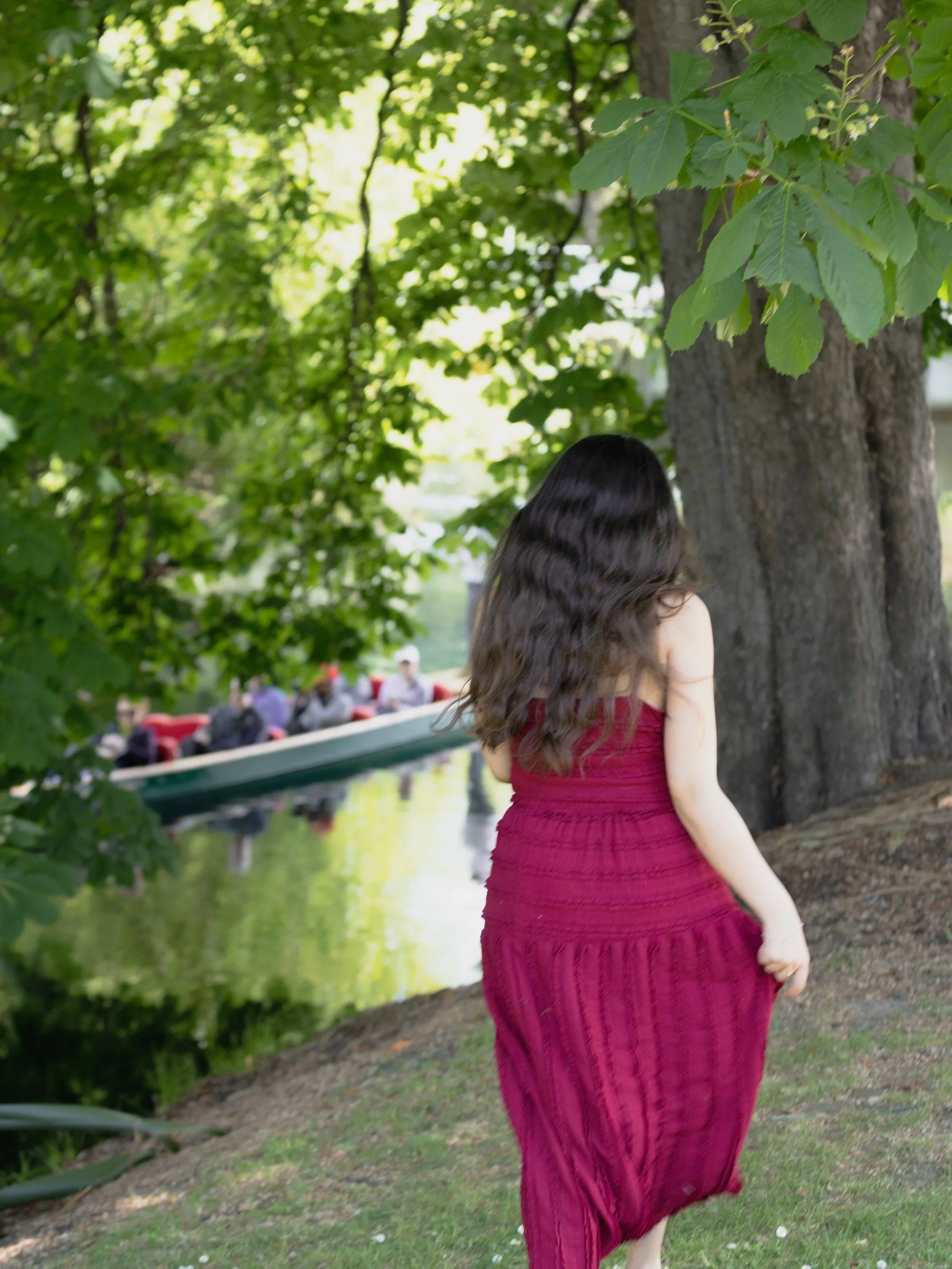 A young girl with long, wavy dark hair wearing a maroon dress walking near a large tree by a river, with a boat and people in the background.