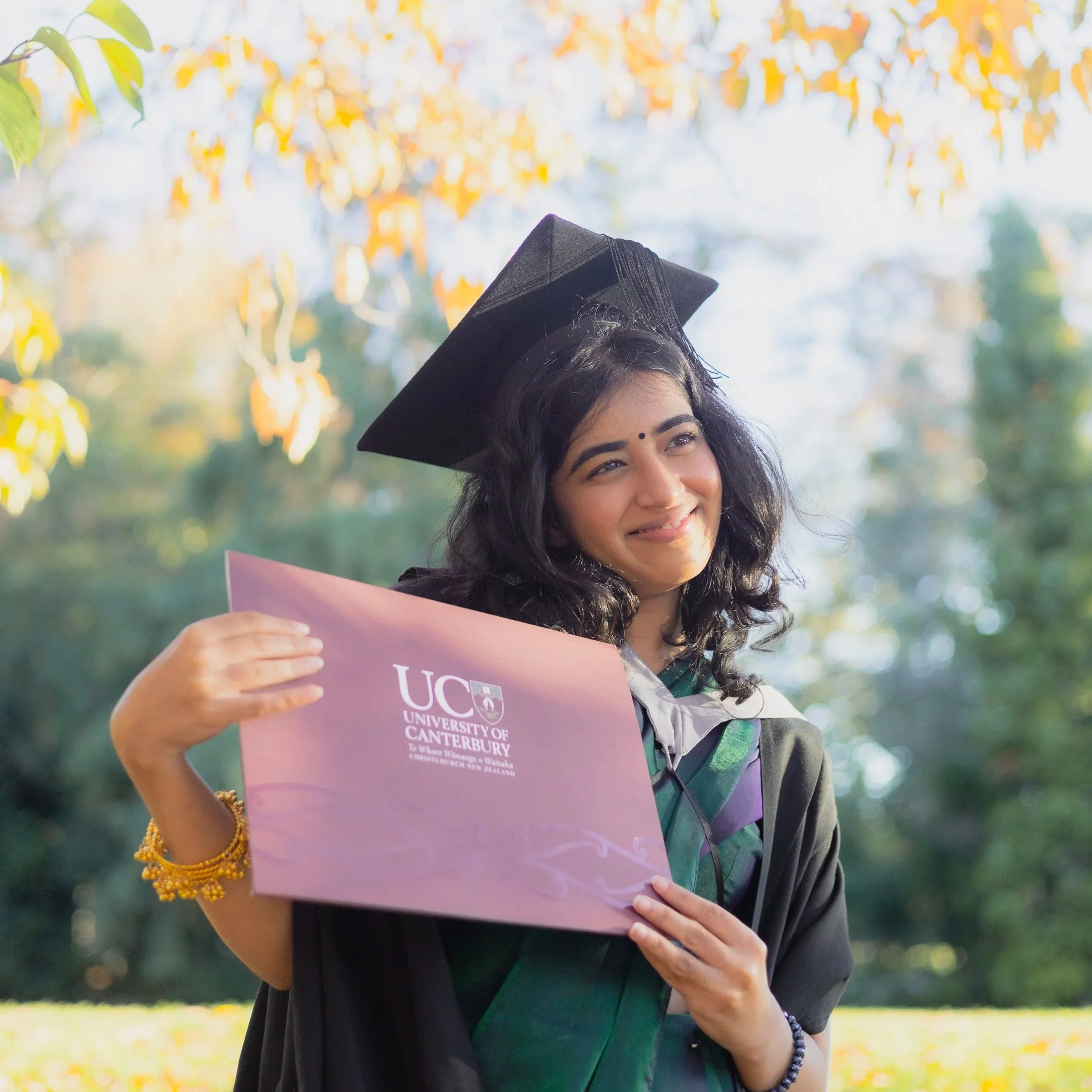 Woman wearing a black graduation cap and gown holding a pink graduation certificate outdoors with green and yellow foliage in the background.