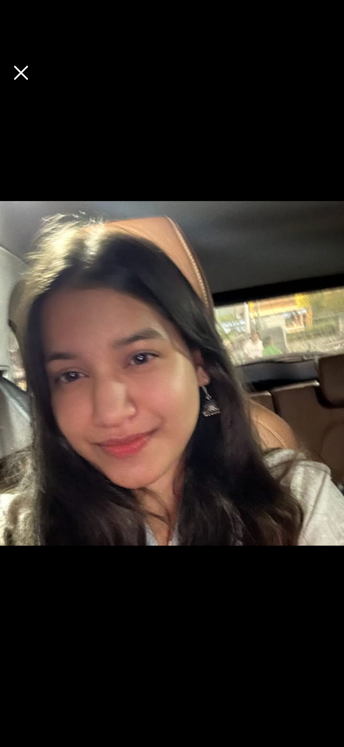 A young woman with long dark hair smiling, seated in a vehicle with tan leather seats, outside view through the window showing buildings and people on the street.
