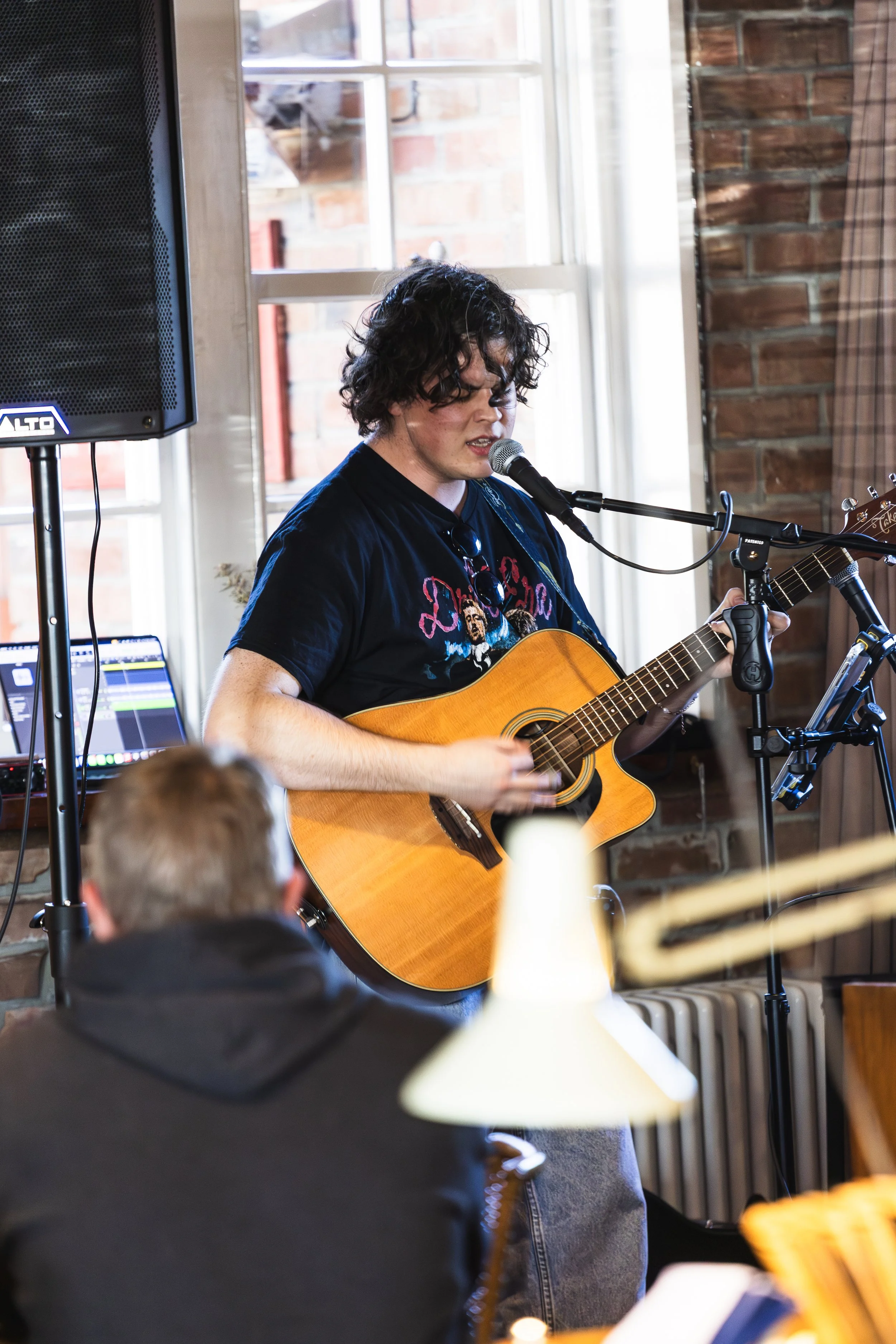 A young man with curly hair is playing an acoustic guitar and singing into a microphone in a cozy indoor setting with brick walls and large windows.
