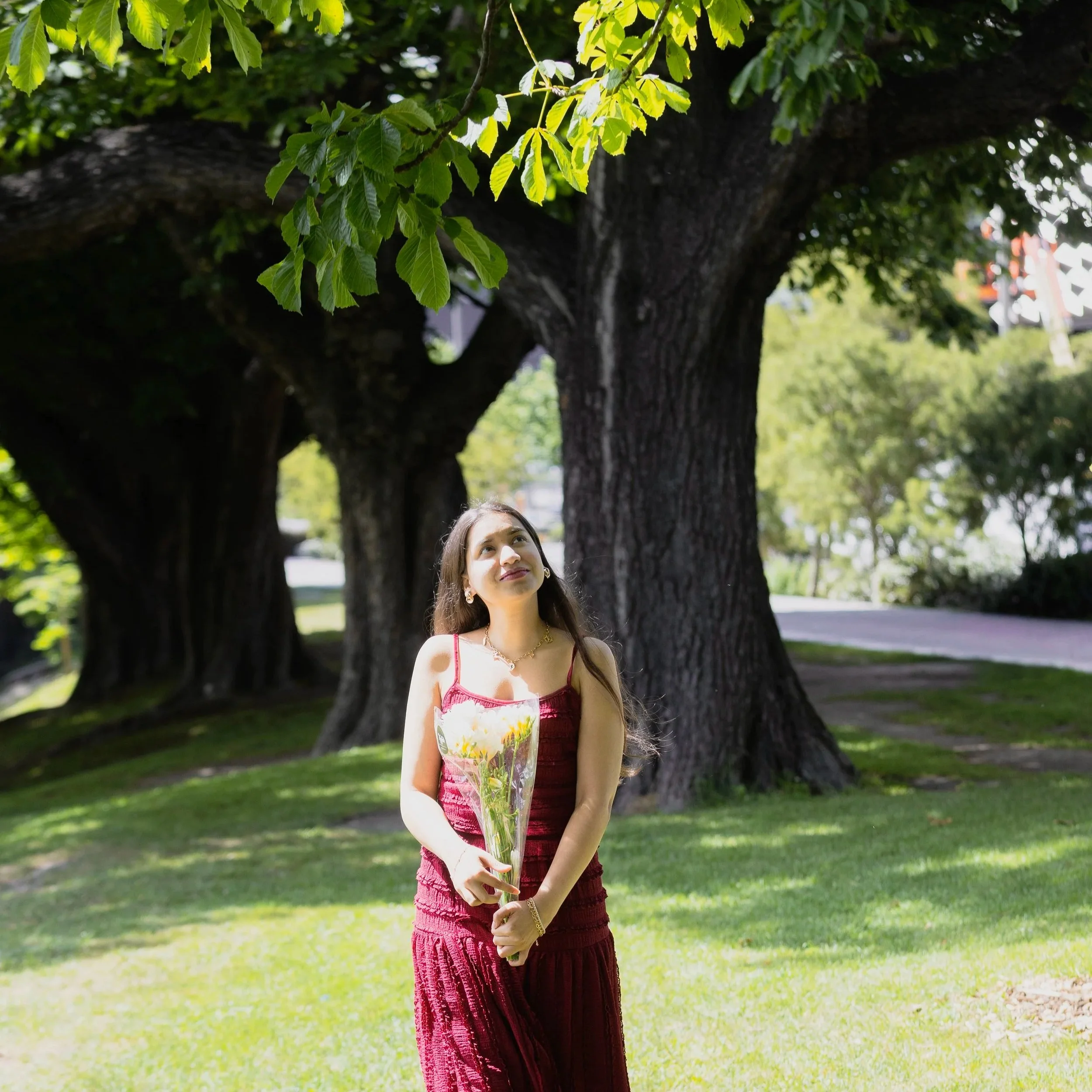 A woman in a red dress holding a bouquet of flowers stands in a park with large trees and green grass, looking up.