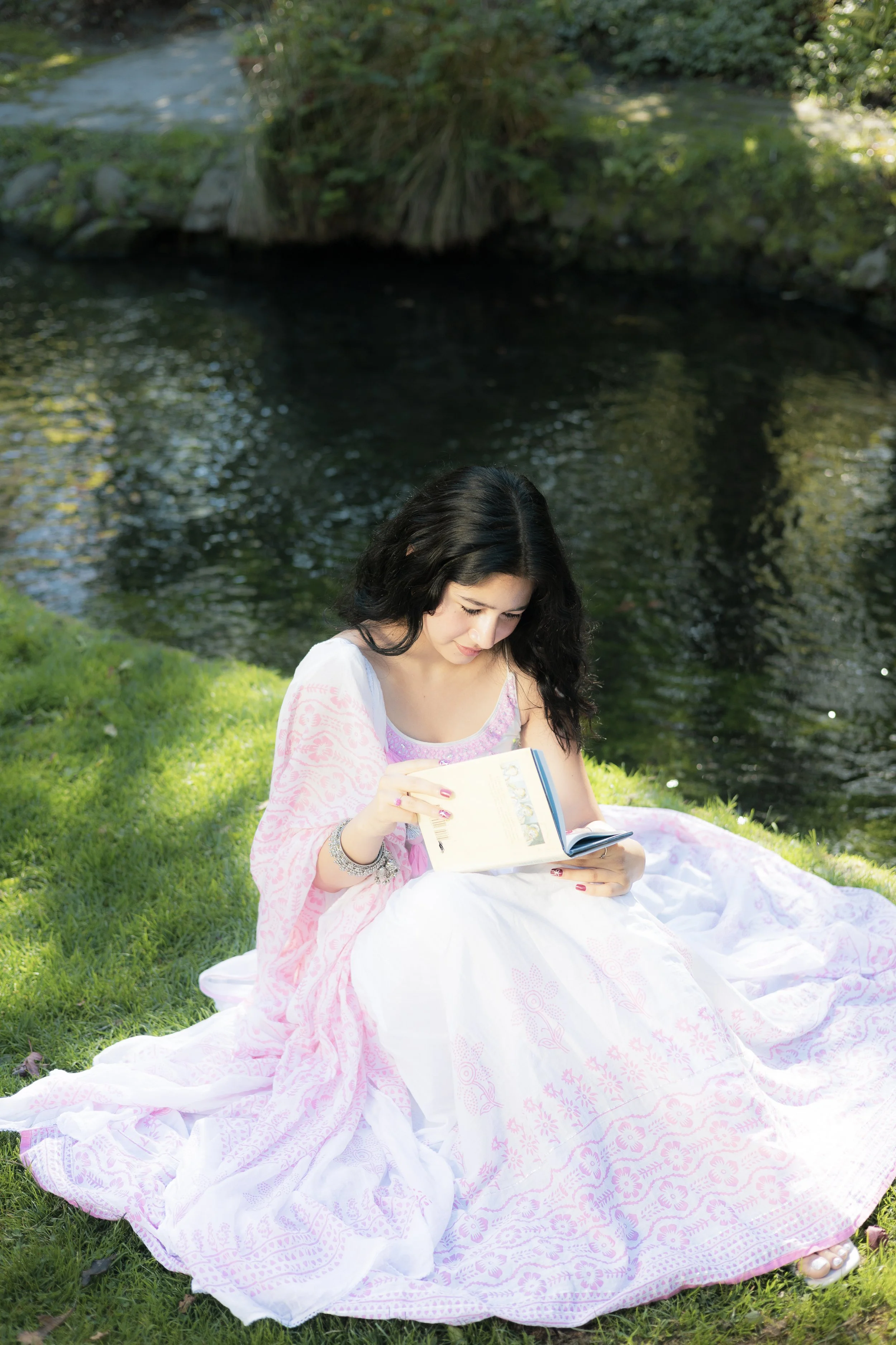 Young woman with black hair reading a book while sitting on a pink and white patterned blanket on the grass near a creek surrounded by greenery.