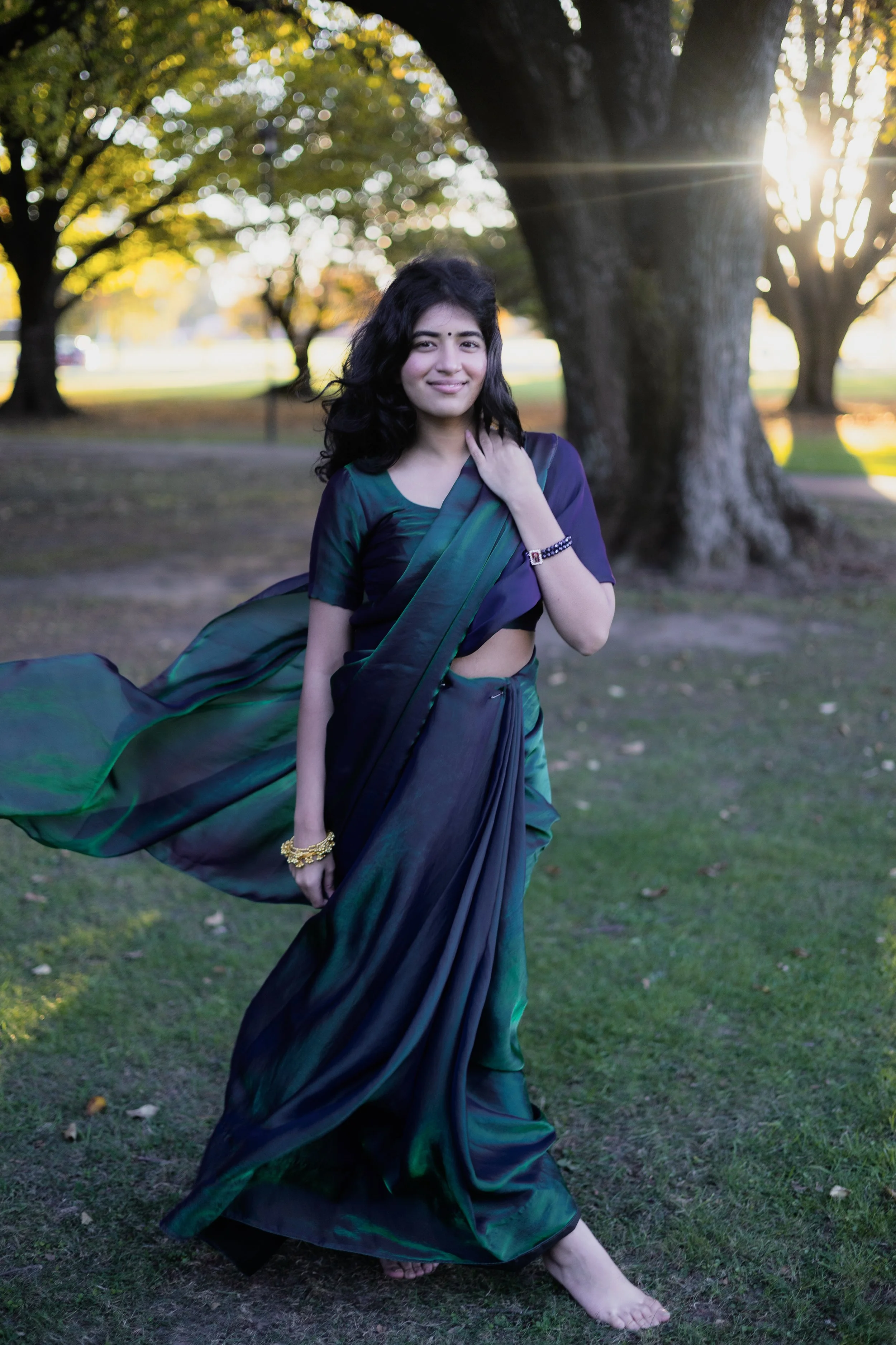 A woman wearing a dark blue and green saree standing outdoors on grass near trees, with sunlight shining through the trees in the background.